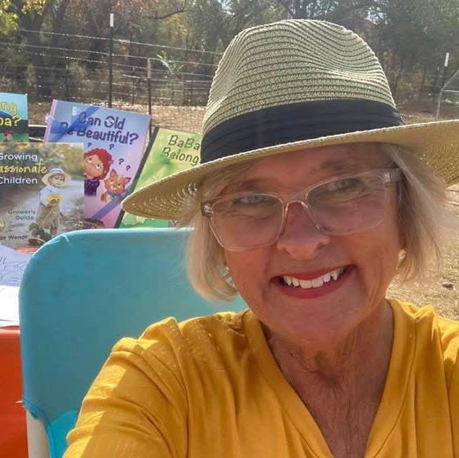 Smiling woman wearing glasses and a straw hat, sitting outdoors with trees and signs in the background.