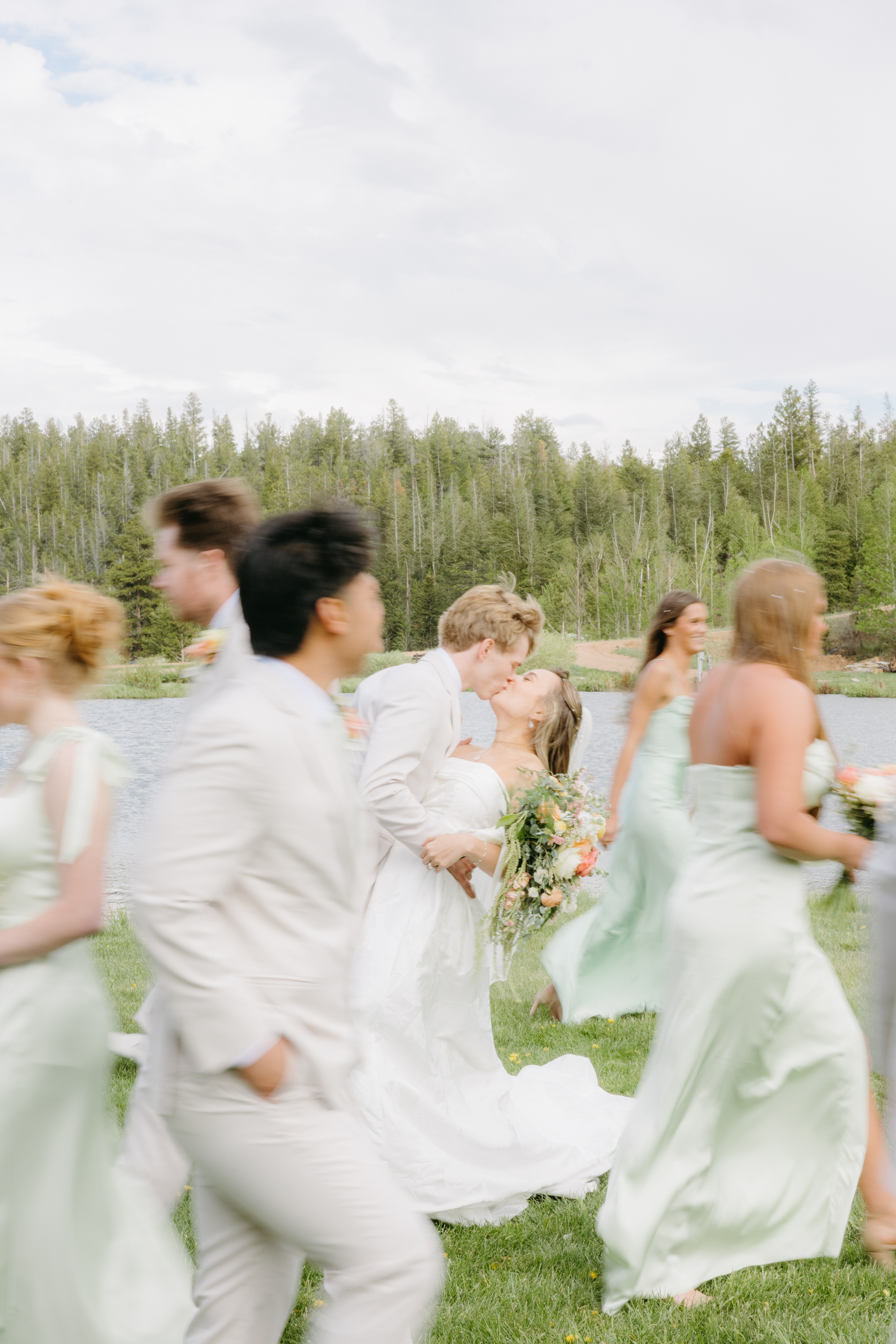 Group of people in light-colored dresses and suits outdoors on grass with trees in background.