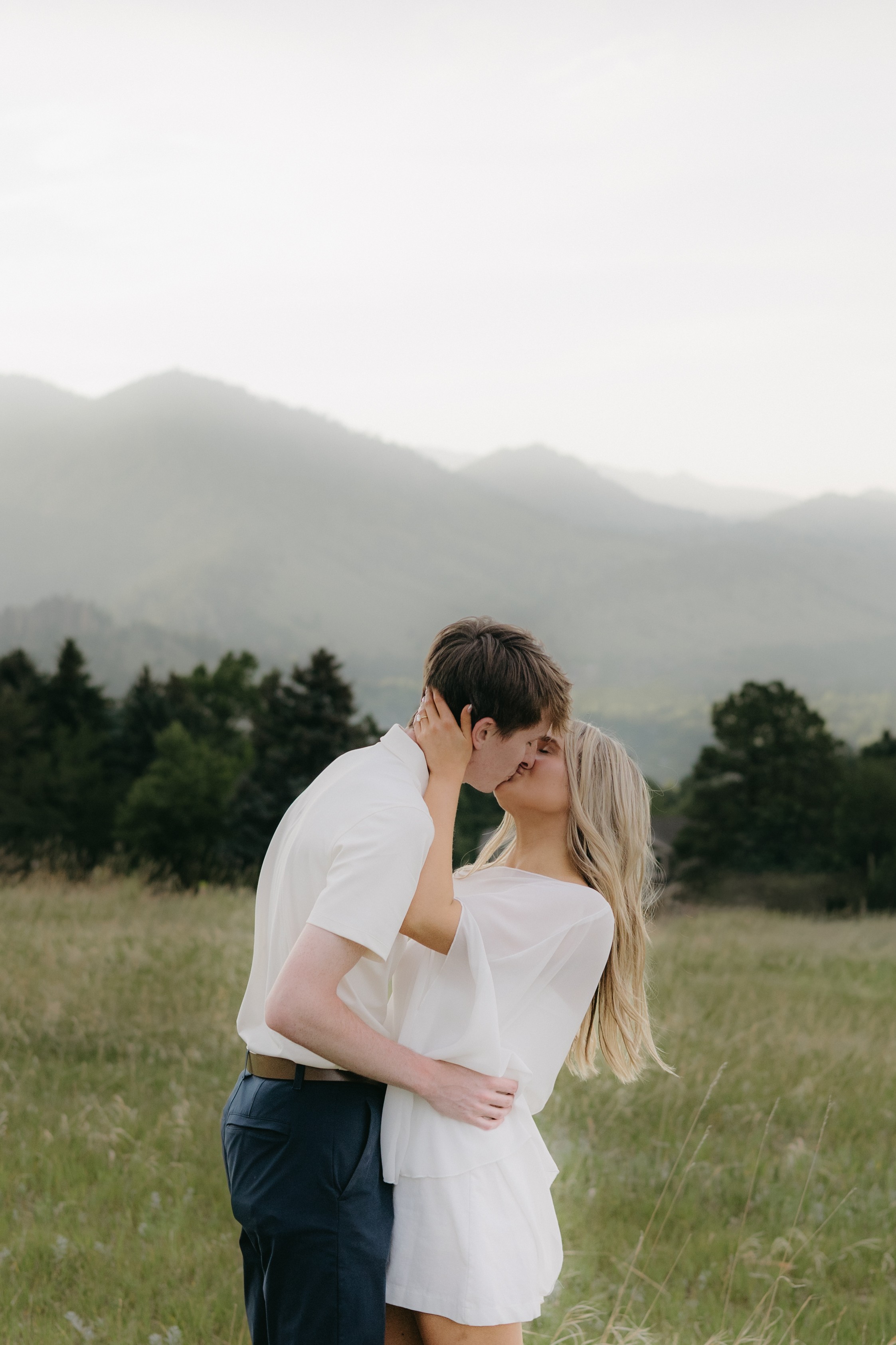 A couple stands in a grassy field, embracing and about to kiss, with mountains and trees in the background.