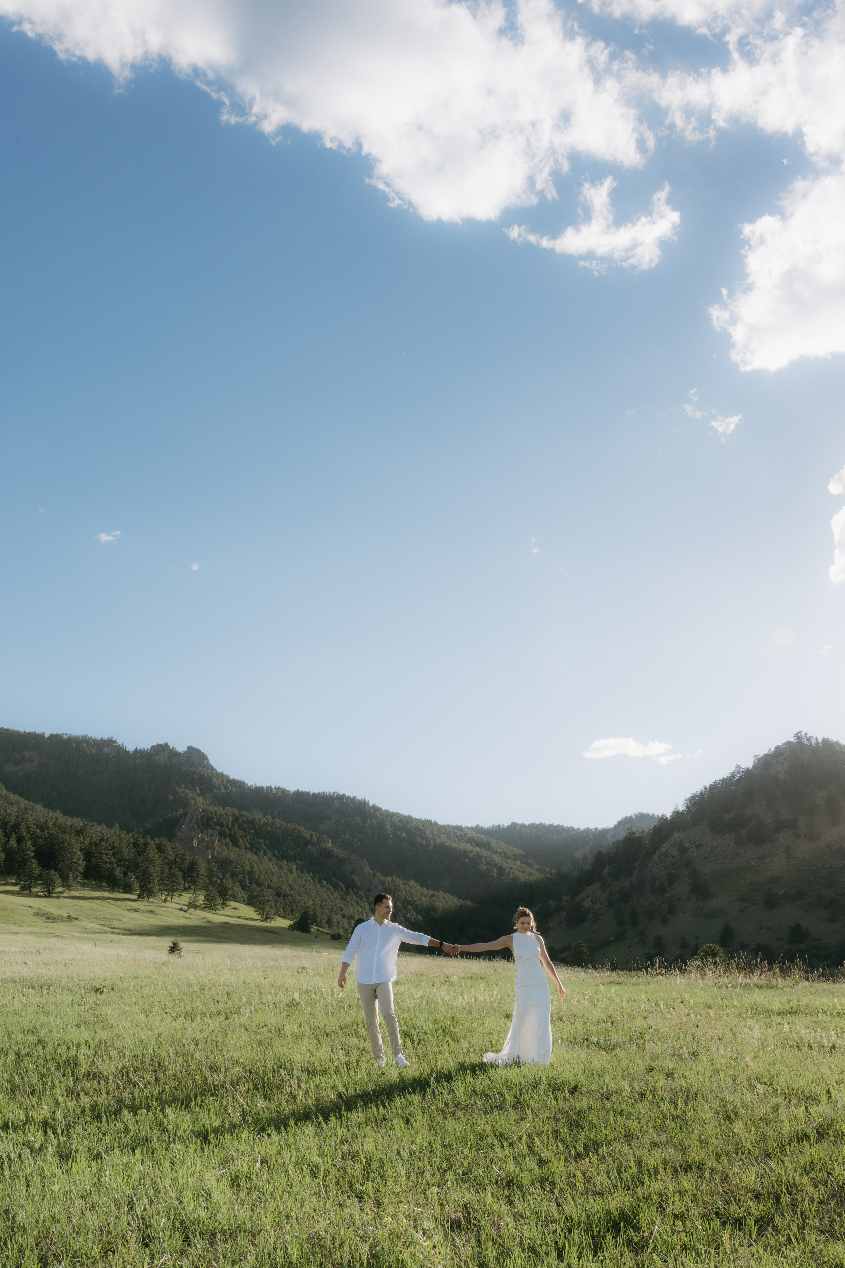 Two people holding hands in a green field with hills and a blue sky with clouds above.