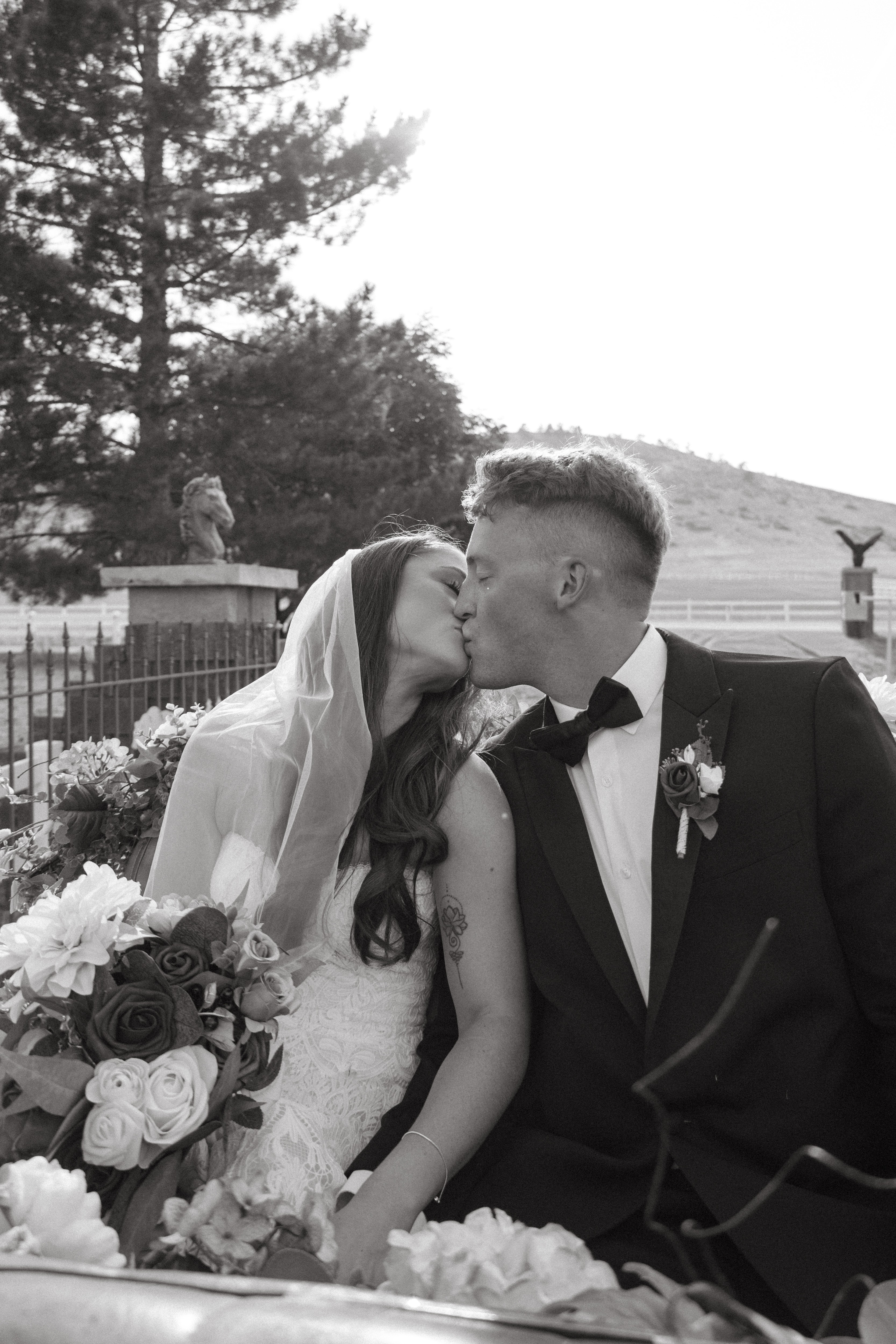 Bride and groom kiss outdoors, bride holding a bouquet of roses, trees and a fence in background.