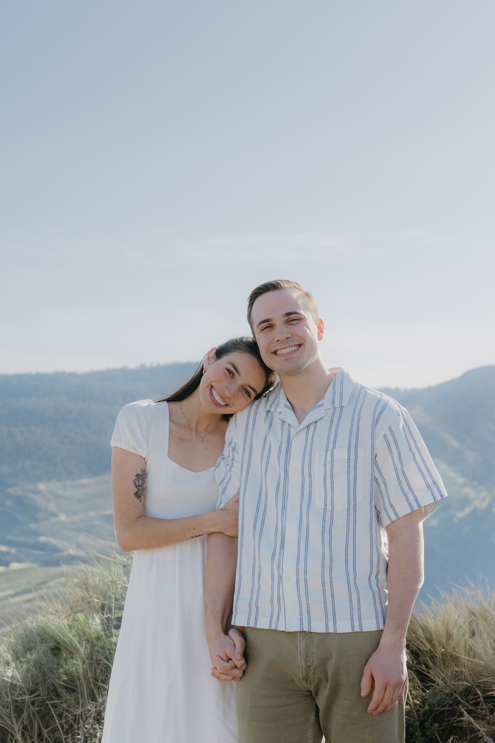 A smiling couple holding hands outdoors with hills and sky in the background.