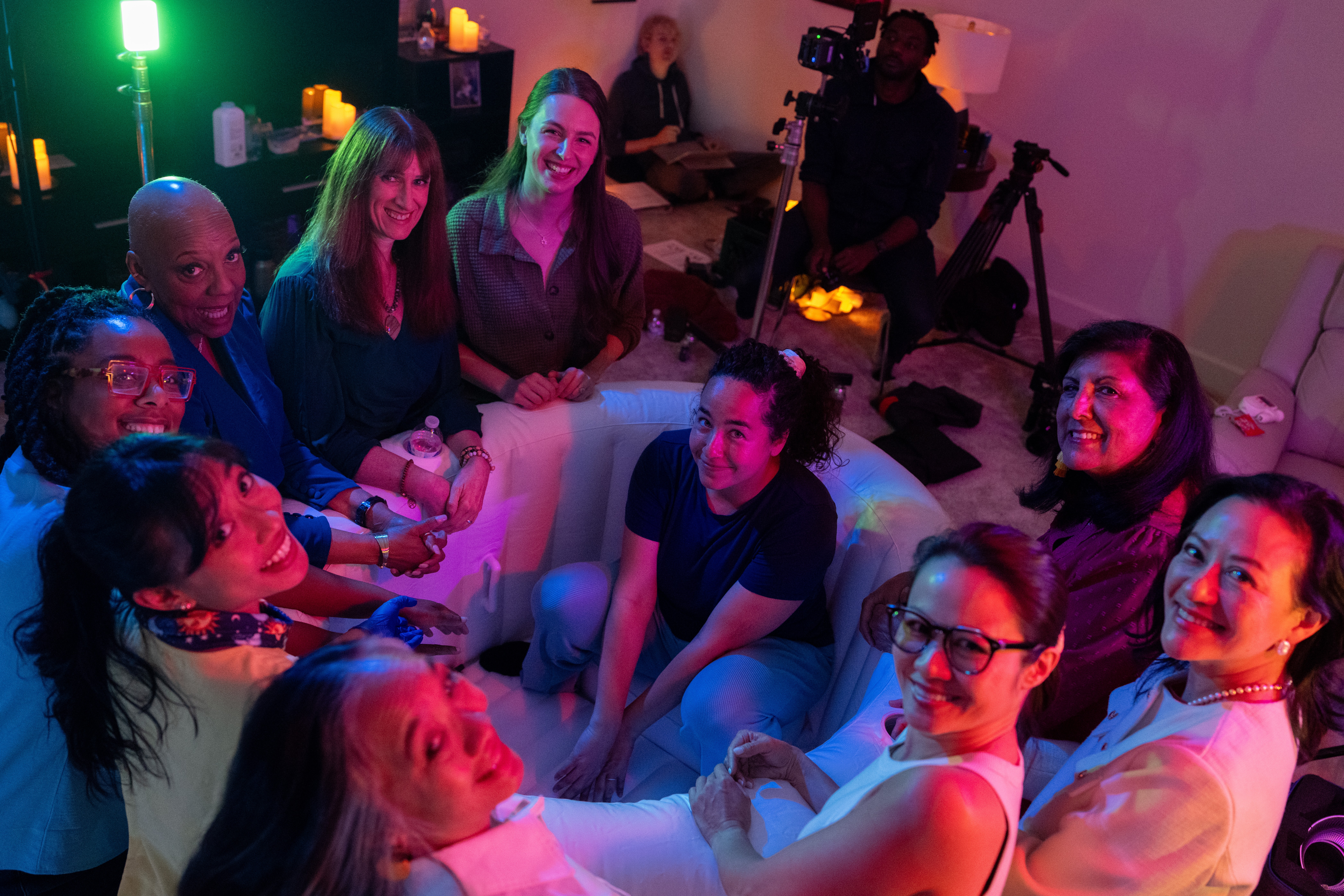 Group of women sitting on couches and chairs in a room with colorful lighting, smiling and talking.