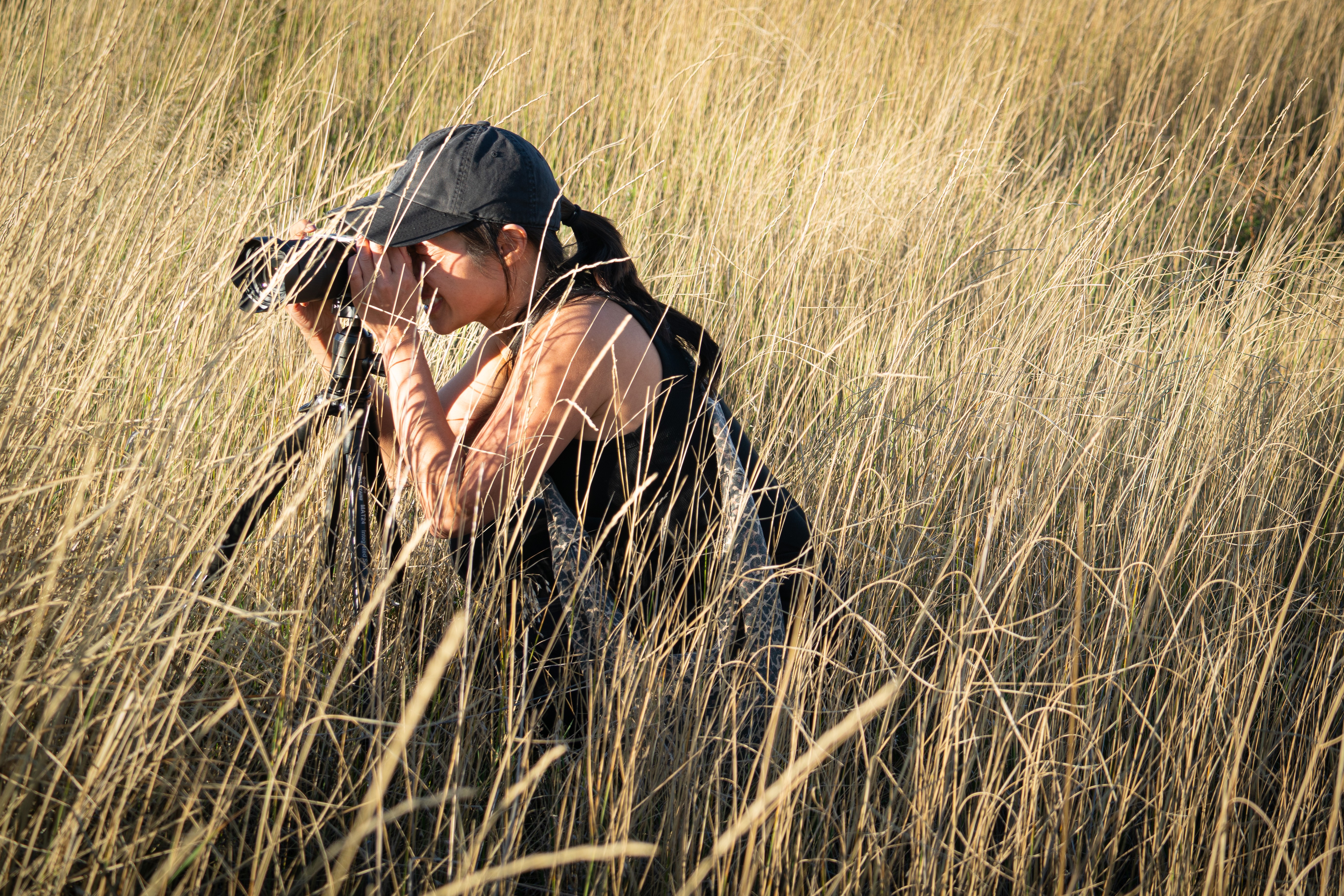 Person crouching in tall grass, wearing a cap and sleeveless shirt, holding binoculars to their eyes, outdoors in a grassy field.