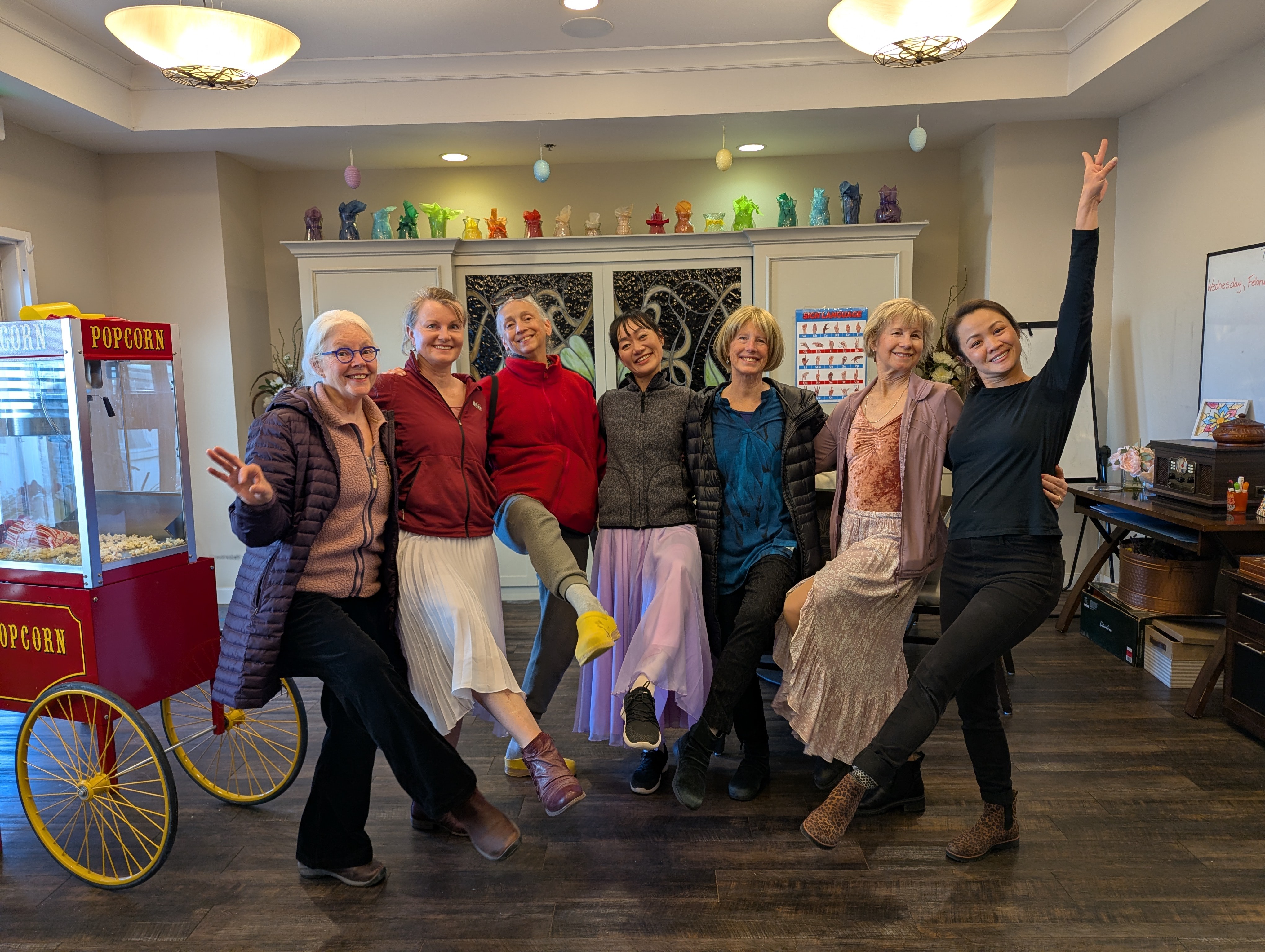Seven women stand together indoors, smiling and posing, with a popcorn machine and colorful decorations in the background.