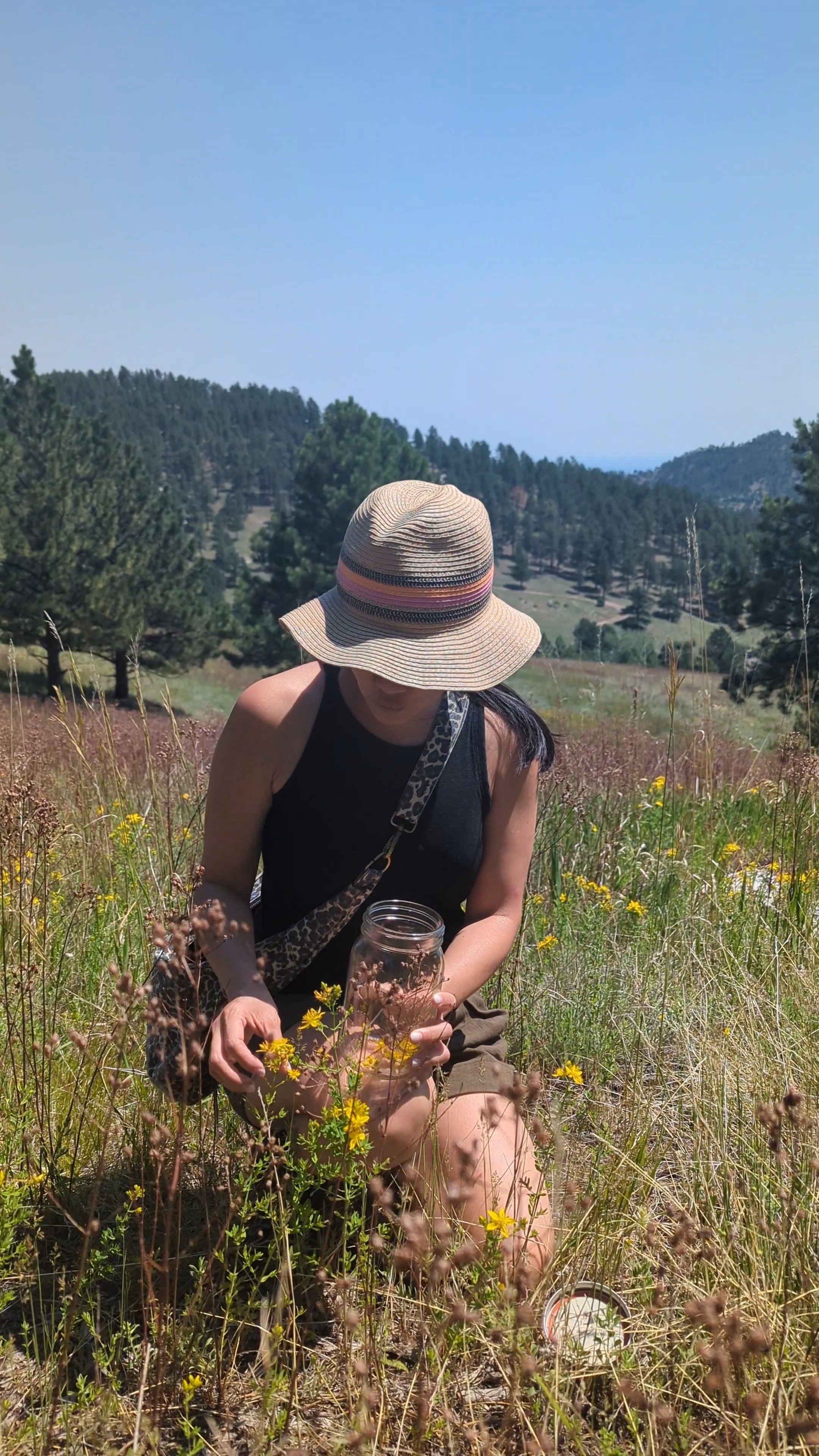 Person sitting in a field with wildflowers, wearing a wide-brimmed hat and black sleeveless top, holding a glass jar.
