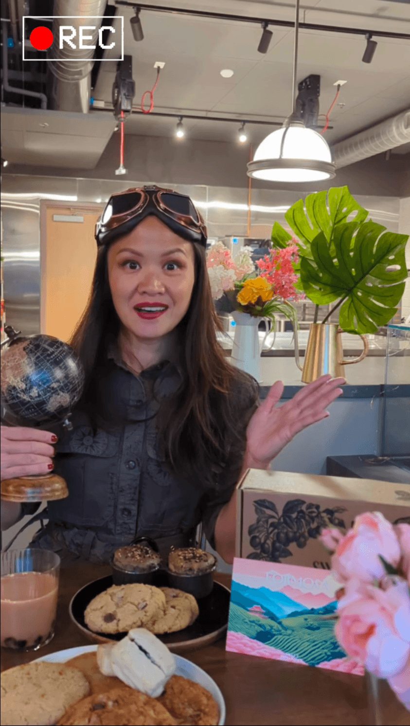 Woman with goggles on her head holding a globe, smiling in a cafe with desserts and a colorful painting nearby.