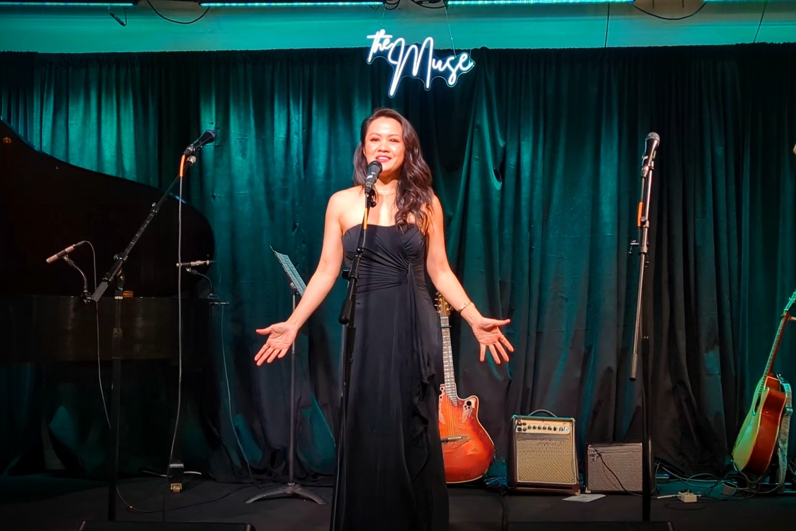 A woman in a black dress performs on stage with a microphone, surrounded by musical instruments and equipment, in front of a teal curtain.
