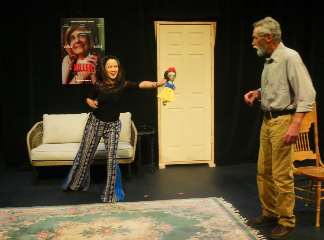 Two people on a stage with a sofa, a door, and a poster, one holding a puppet, the other smiling.