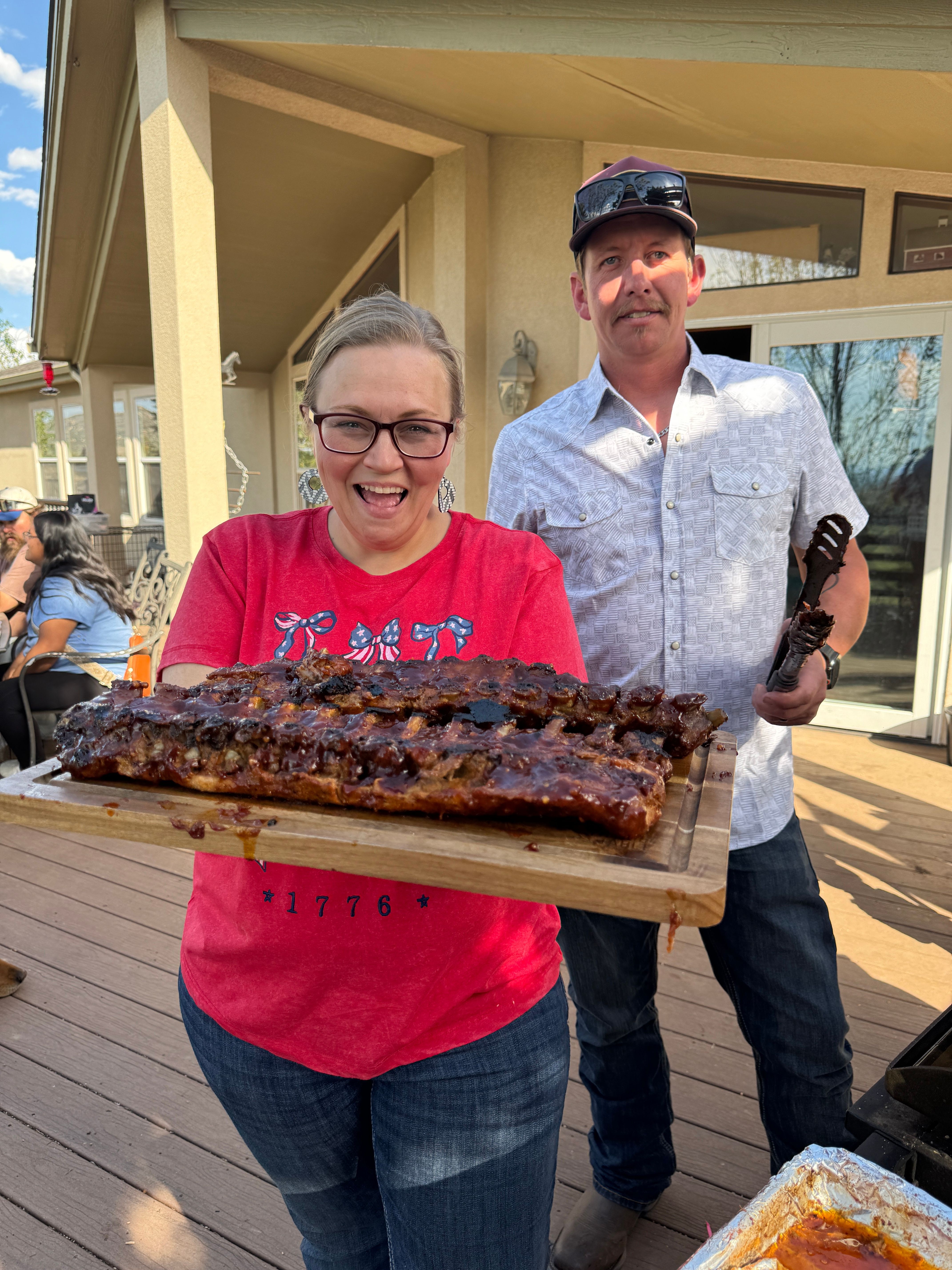 A woman in a red shirt and glasses holds a large tray with a long, cooked meat dish. A man in a white shirt stands nearby.