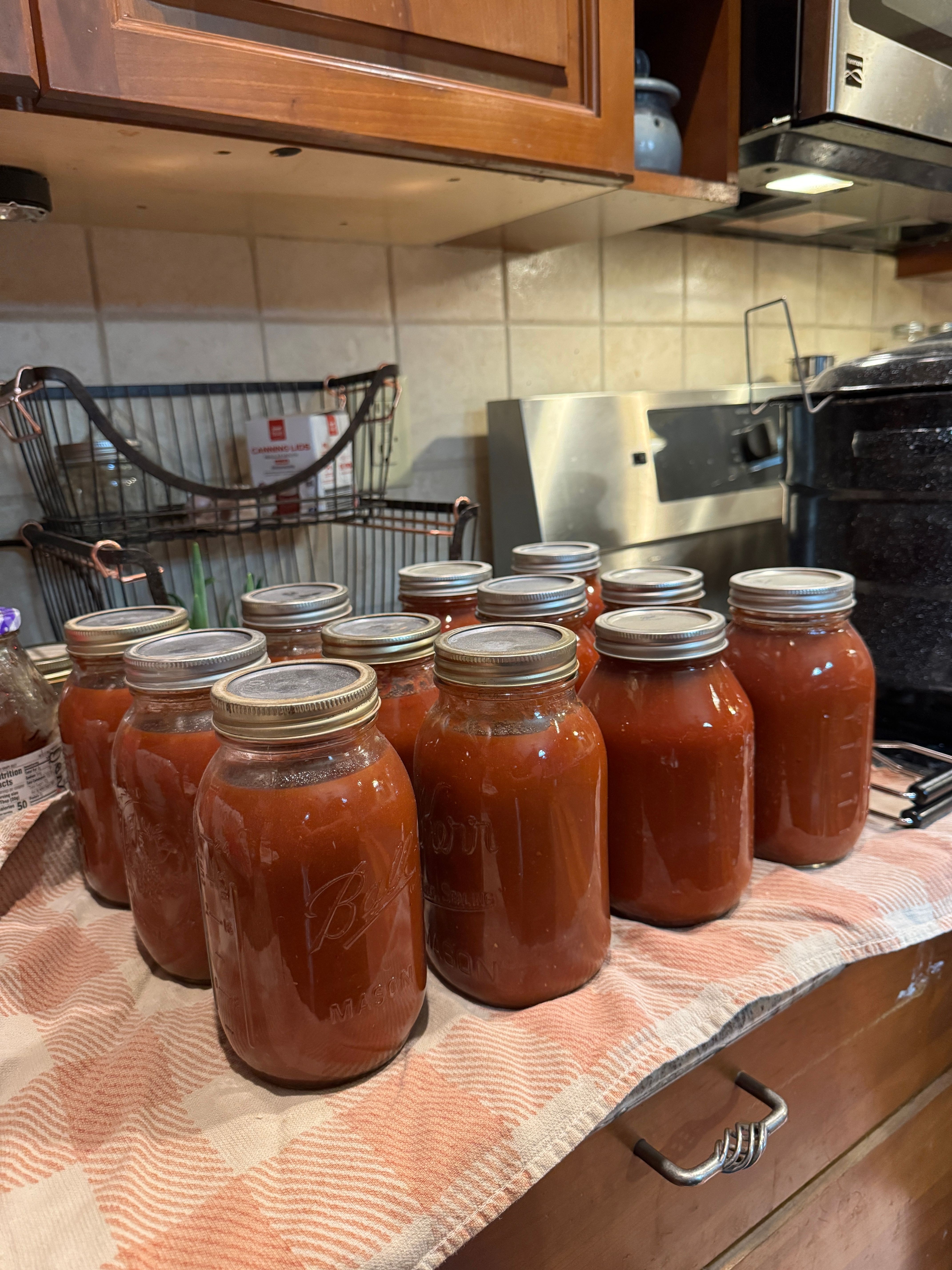 Seven jars filled with a red sauce or jam are arranged on a tablecloth-covered surface in a kitchen. The jars have metal lids.