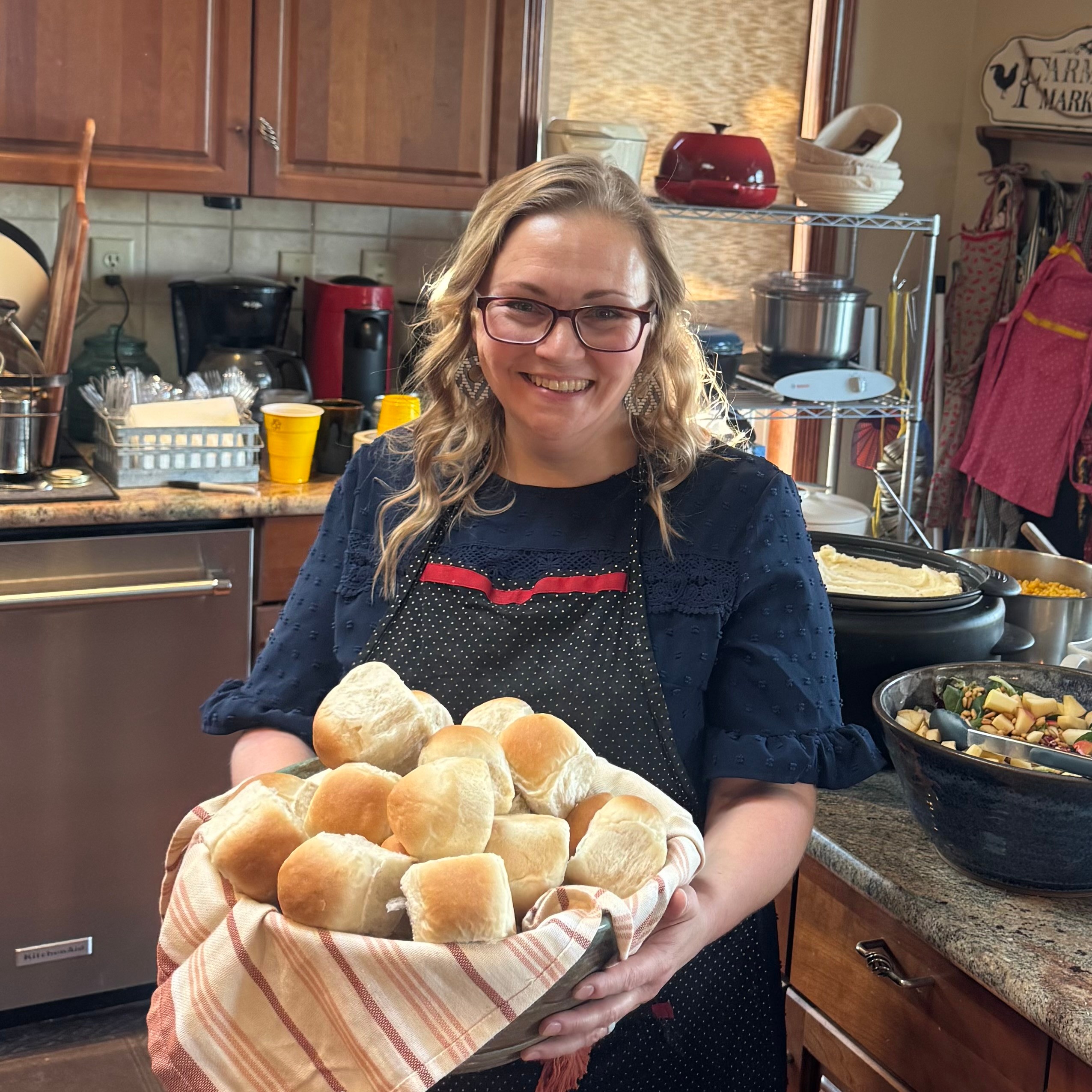 Woman with glasses and wavy hair holding a basket of bread rolls in a kitchen.