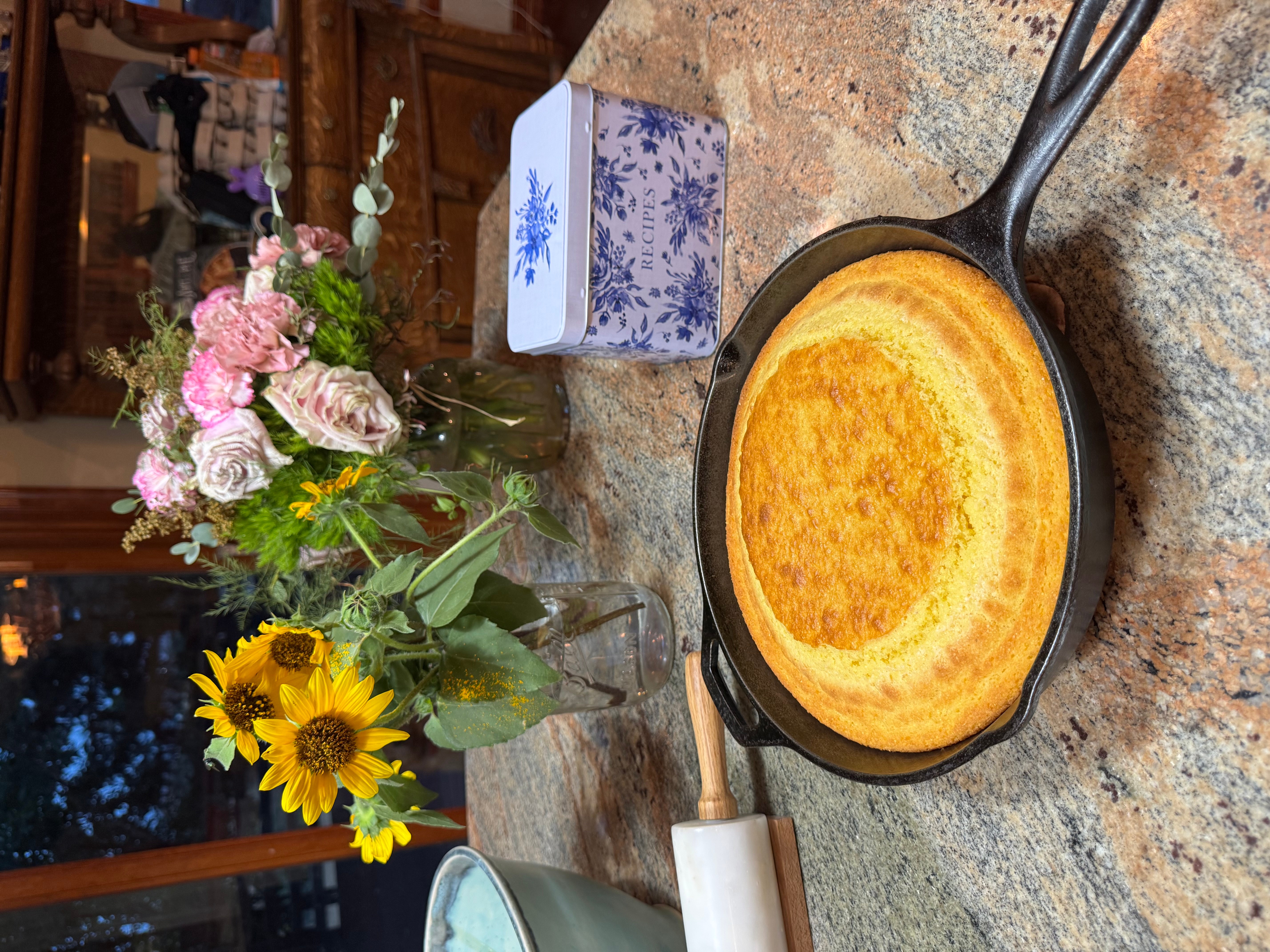 Fried dish in a skillet, flowers in a jar, a tissue box, and a wooden-handled utensil on a kitchen counter.
