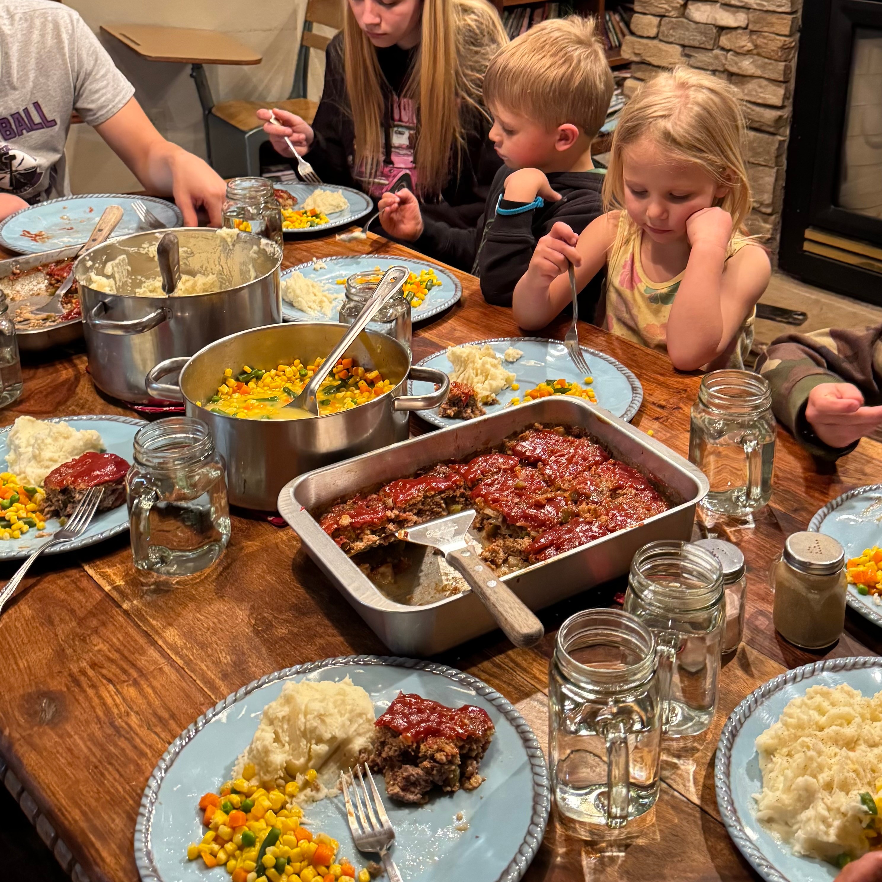 Children and adults sit around a table with food, including casseroles, mashed potatoes, and drinks, in a cozy setting.