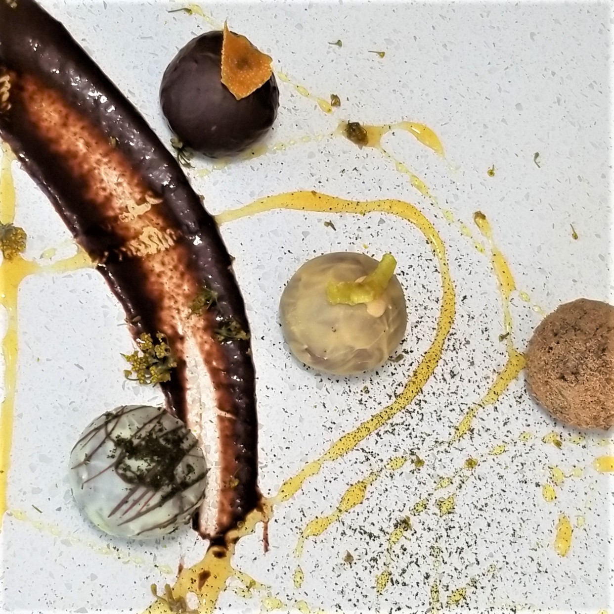 Close-up of a dessert with a chocolate-covered item, a berry, and various garnishes on a white surface, with yellow sauce drizzles.