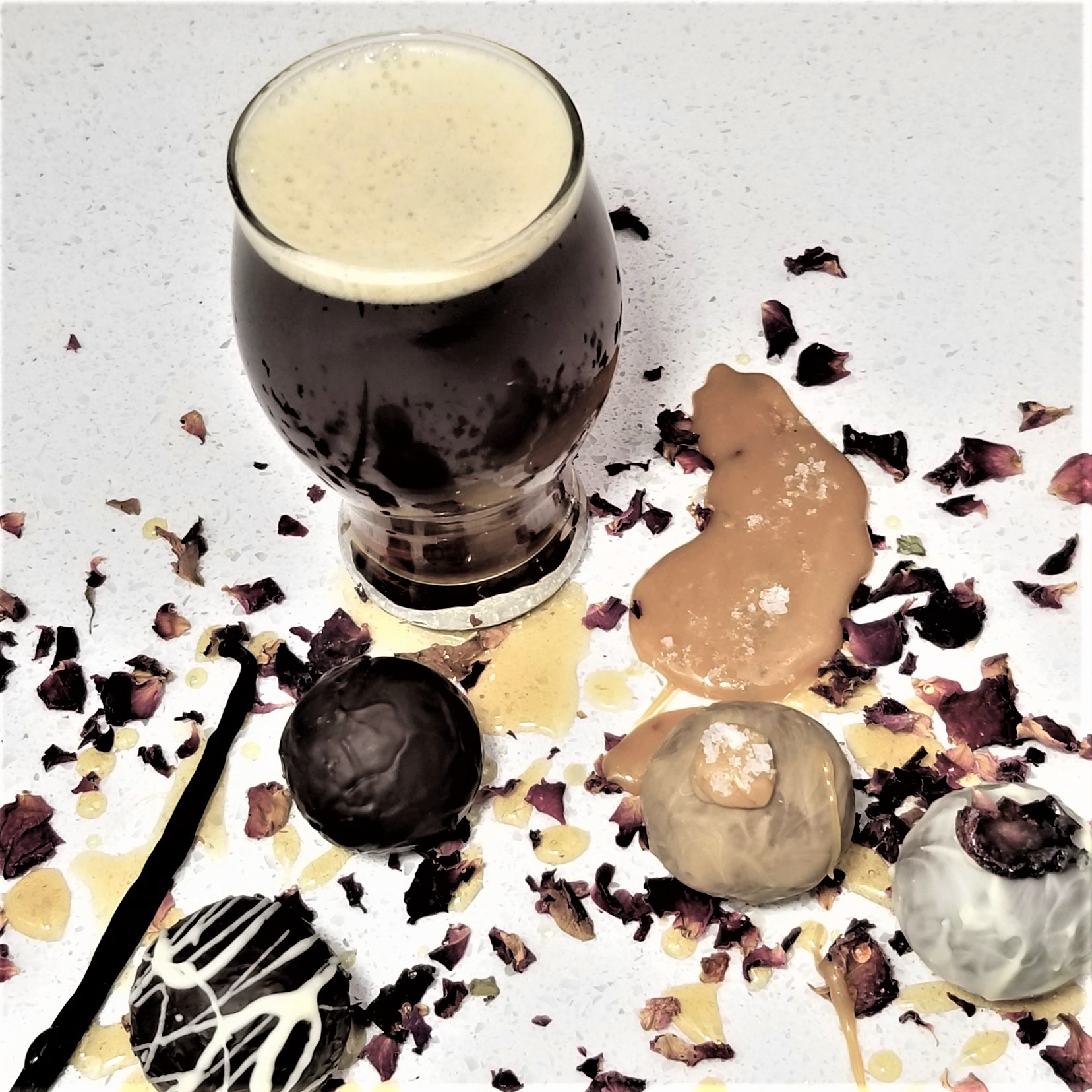 A glass of dark beer with foam, surrounded by ice cream scoops, chocolate, and flower petals on a white surface.