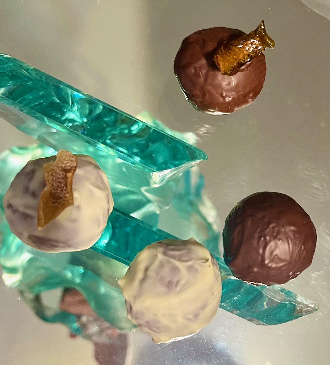 Three chocolate truffles with gold leaf decorations on a reflective surface, with a glass object nearby.