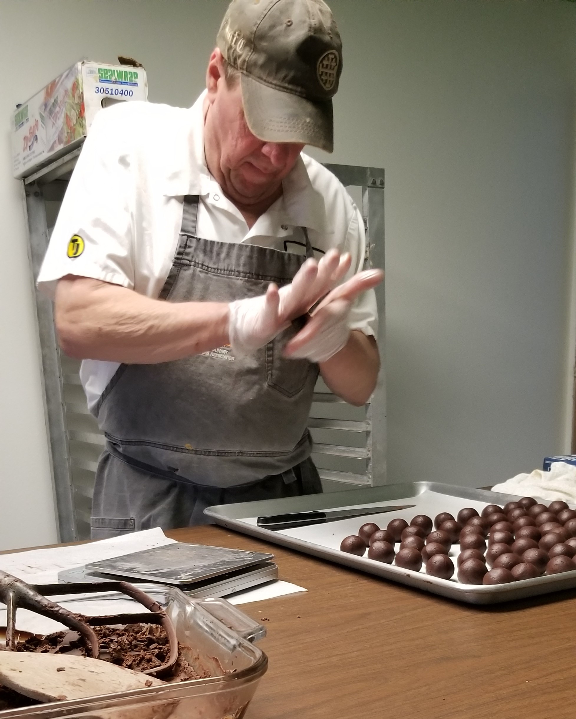 Person wearing a cap and apron, preparing chocolate truffles in a kitchen, with a tray of chocolates on the table.