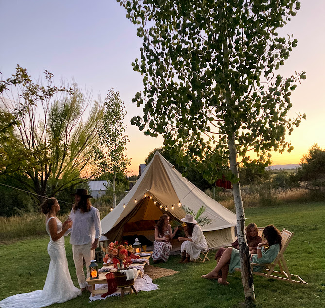 People gather around a tent outdoors during sunset, with trees and grass in the background, some sitting and others standing.
