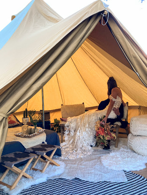 Person in a wedding dress inside a tent with furniture and floral arrangements, sunlight outside, cozy setting.