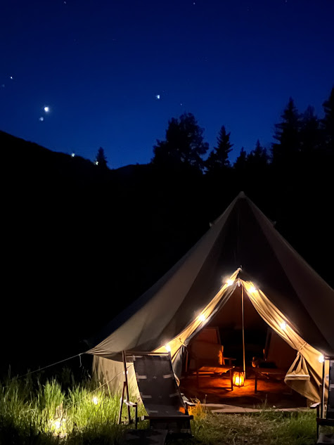 Night sky with stars above a tent, illuminated inside, surrounded by trees and grass, with a camp chair outside.