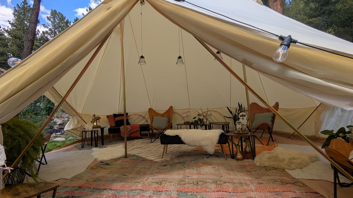 Interior of a large tent with chairs, cushions, and a rug, set in a forested area.