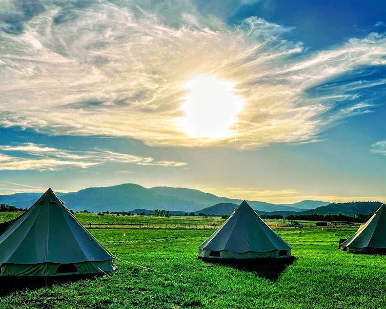 Three tents on green grass with mountains and a bright sun in a partly cloudy sky.