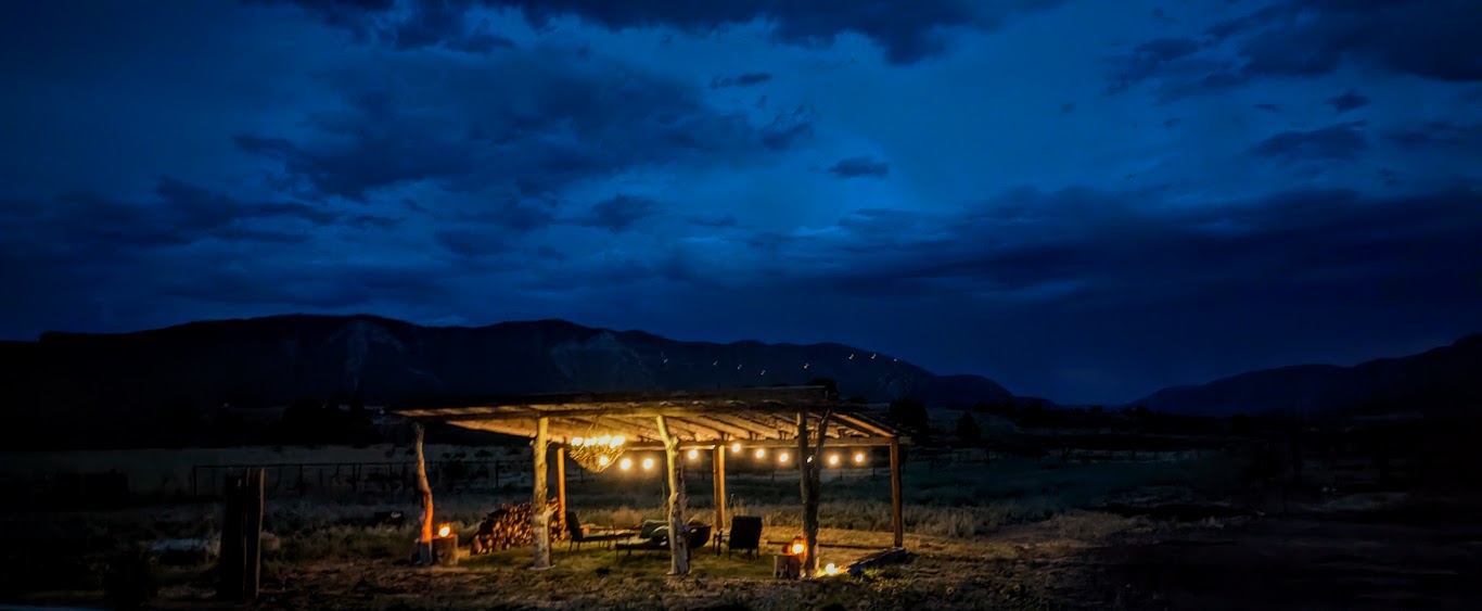 Nighttime scene with a lit wooden shelter in an open field, dark sky with clouds, distant hills, and no people visible.