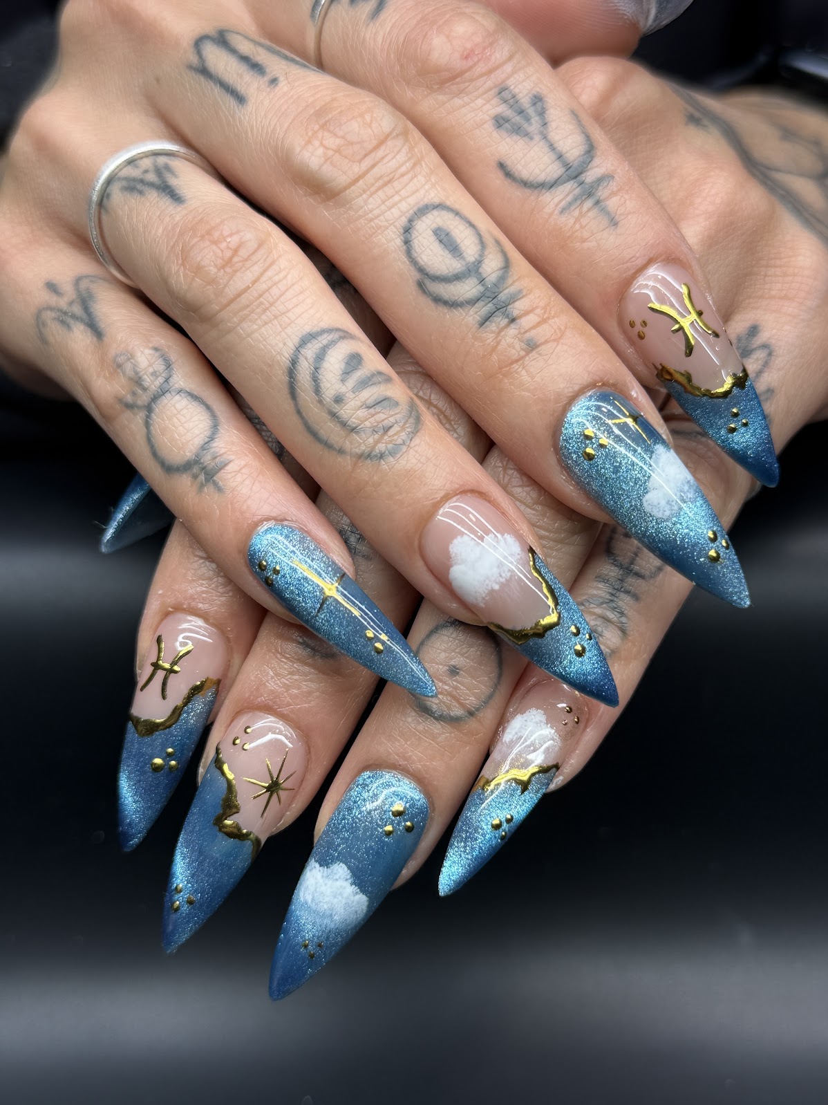 Hands with long, pointed nails painted blue and white, decorated with gold stars and celestial designs, tattoos visible on fingers