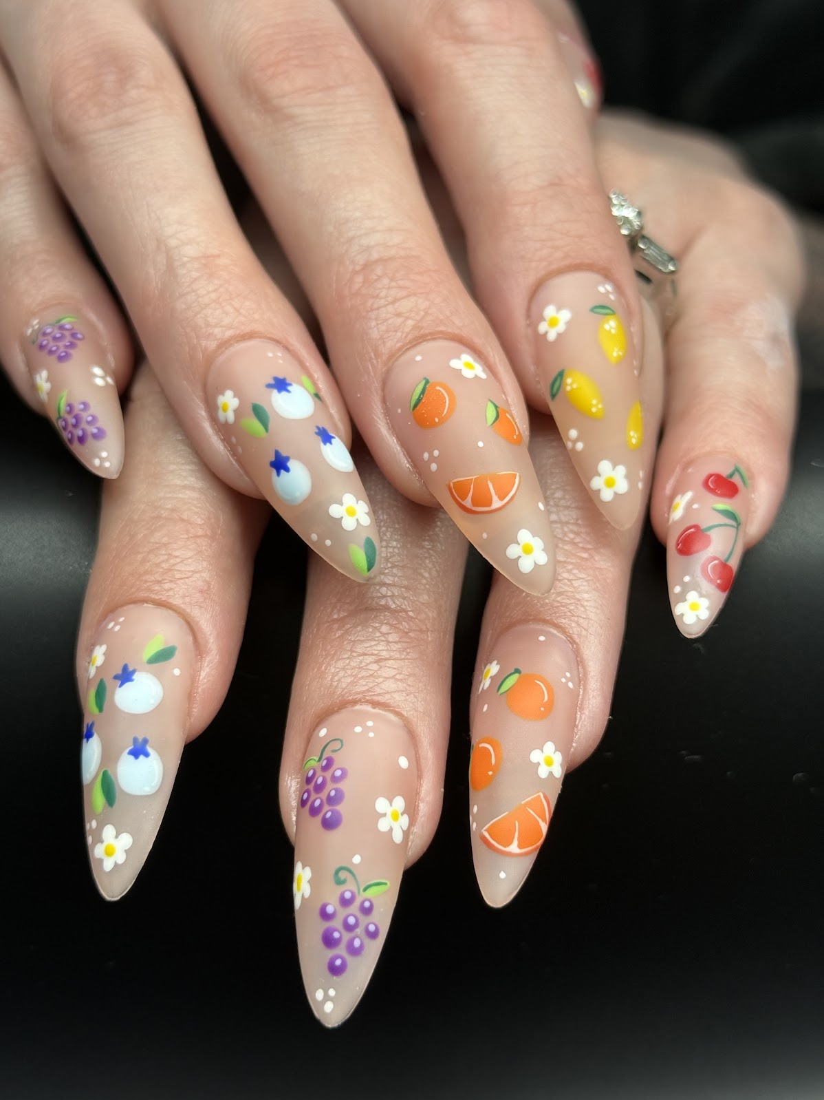 Nail art with colorful fruit and flower designs on long, almond-shaped nails, featuring oranges, grapes, and daisies.