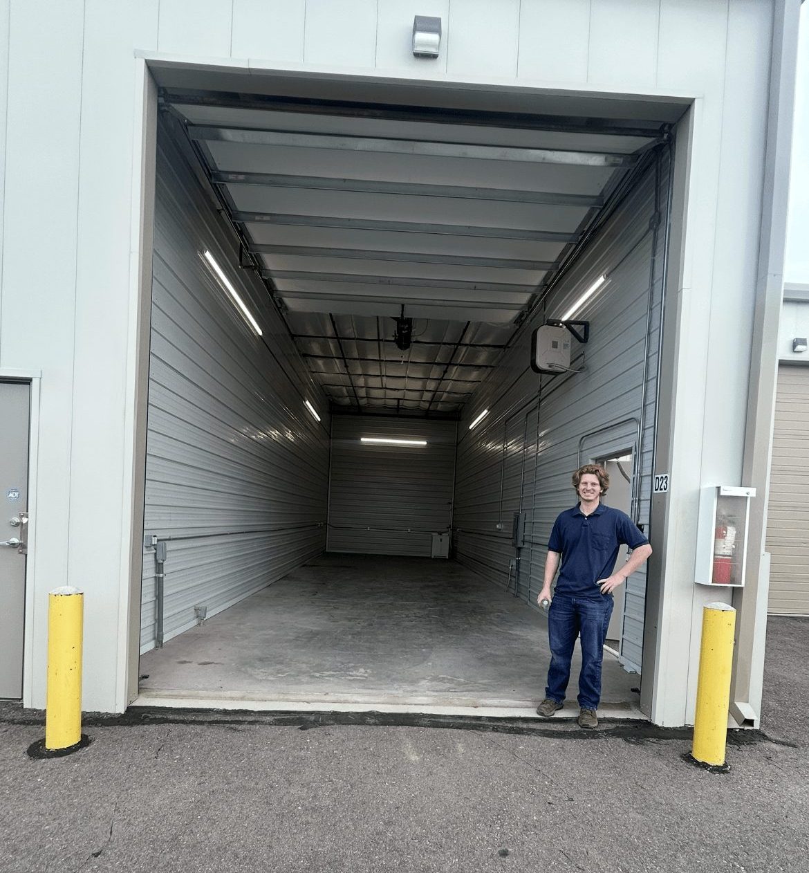 Person standing outside a large industrial loading dock with yellow safety bollards, gray walls, and a high ceiling.