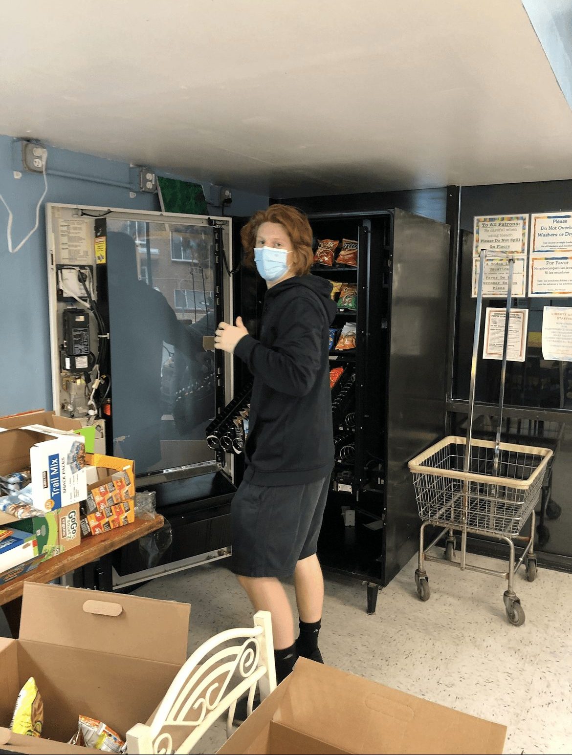Person wearing a mask standing next to a vending machine in a room with carts and boxes.