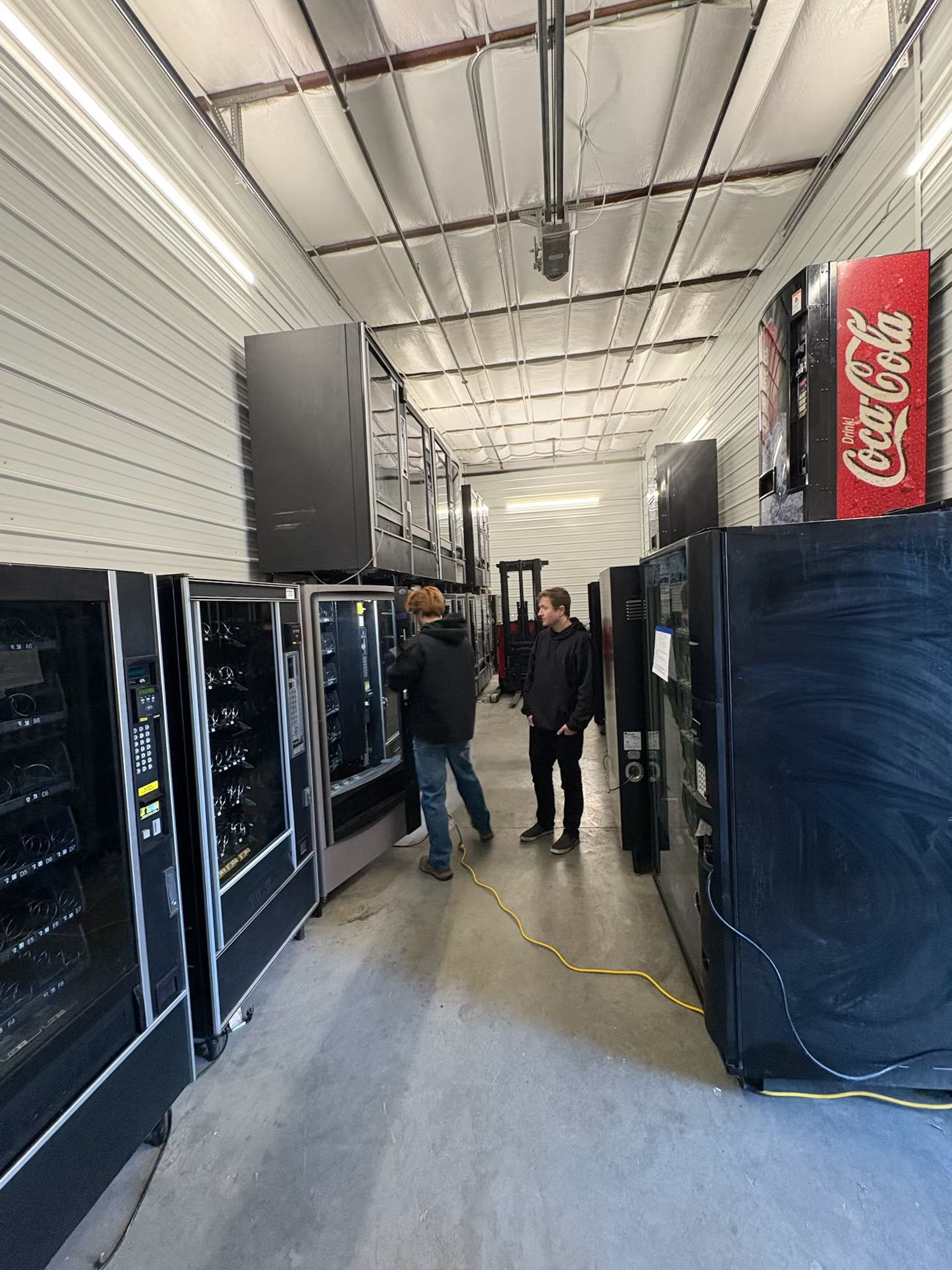 People standing in a warehouse-like space with vending machines and a Coca-Cola sign, viewed from the side.