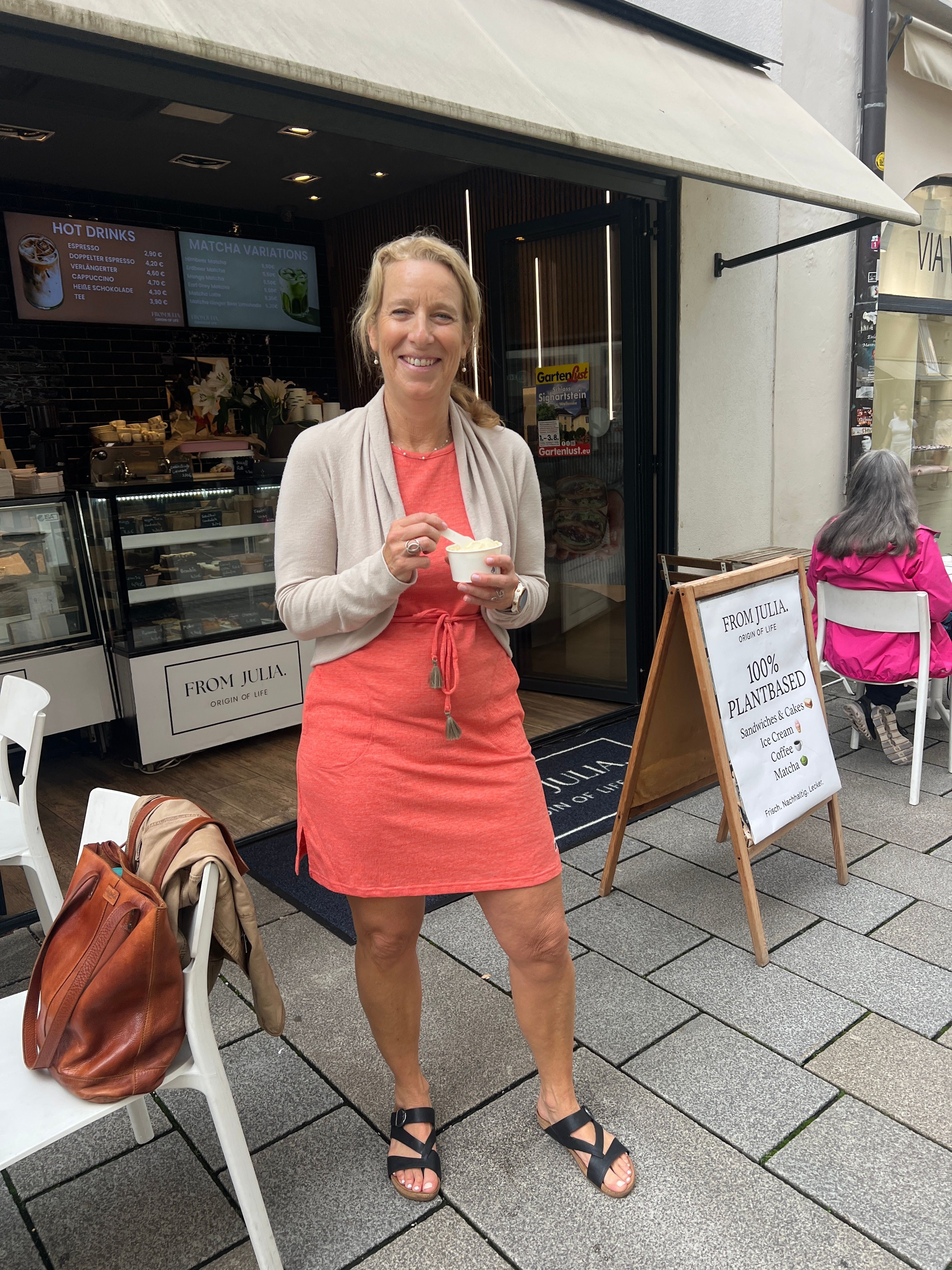 Woman in coral dress and white blazer holding a drink outside a cafe, with a pink bag and a signboard nearby.