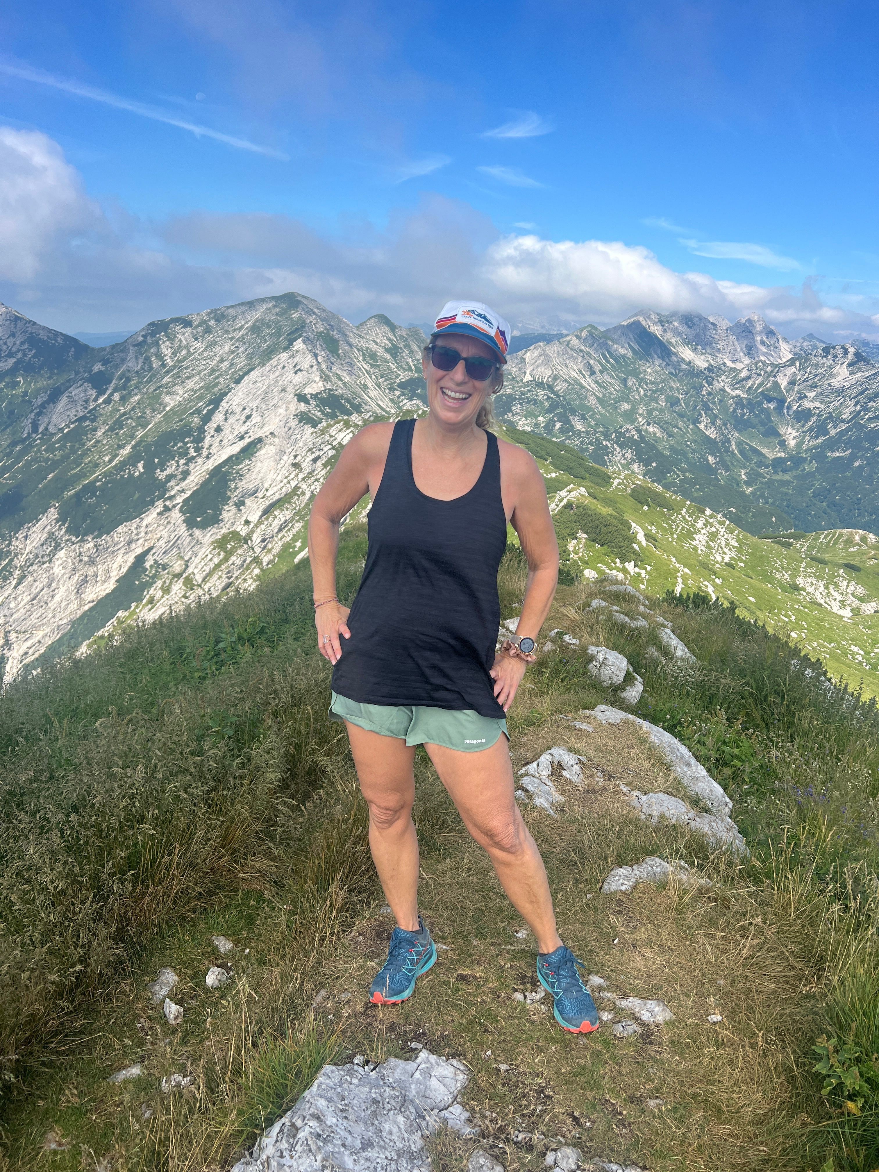 Person standing on rocky mountain trail with green hills and blue sky in background.
