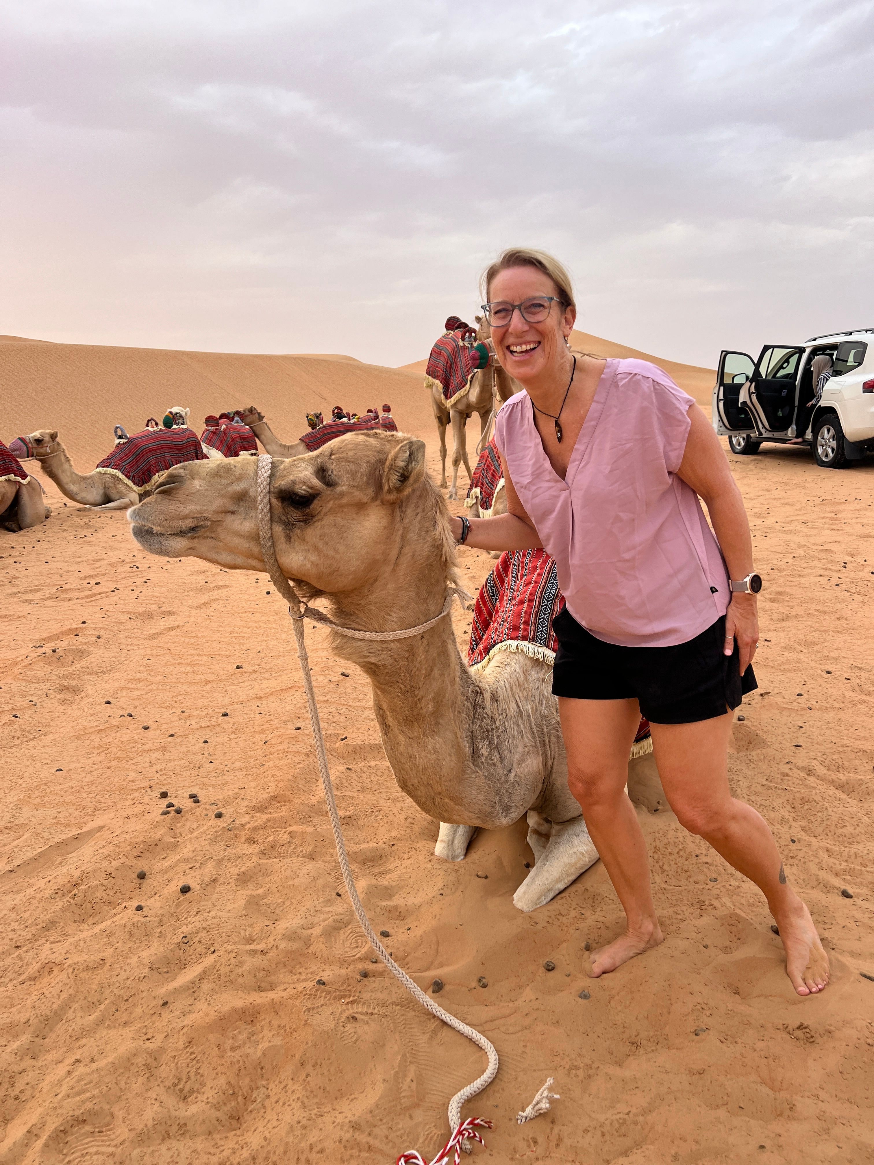 Woman standing on sandy ground holding a leash attached to a dog, with vehicles and a cloudy sky in the background.