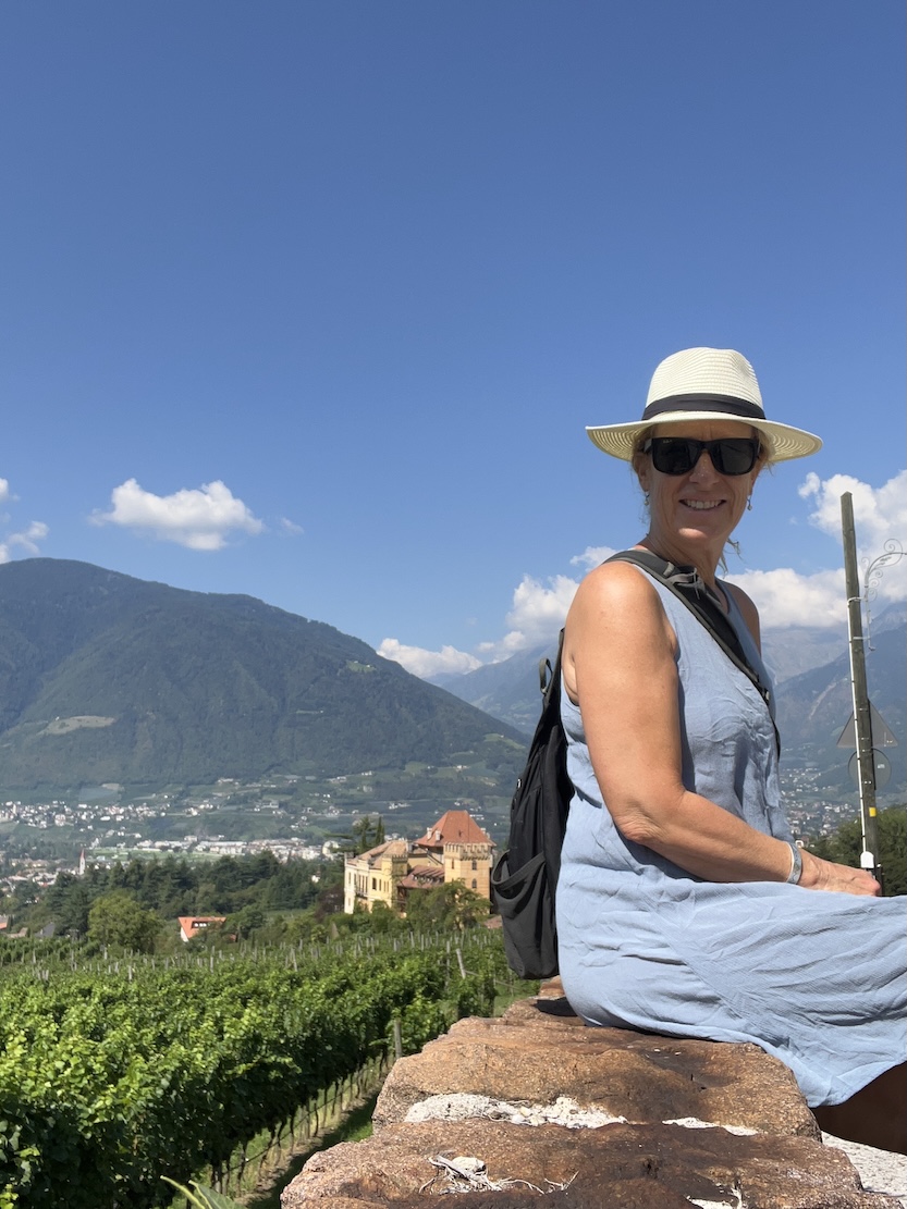 Woman sitting outdoors on a stone surface, wearing a hat and sunglasses, with mountains and a blue sky in the background.