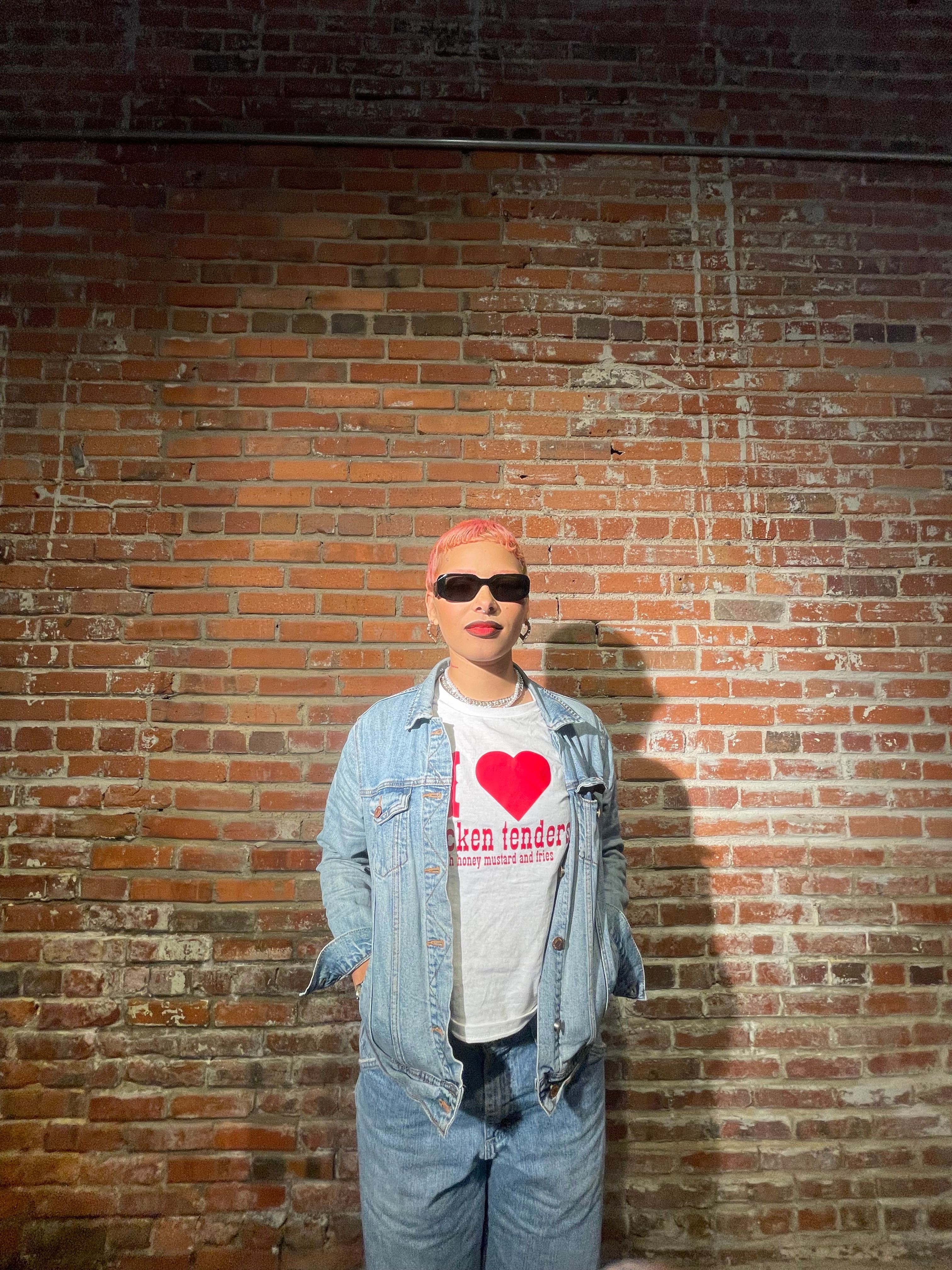 Person wearing sunglasses, a white shirt with a red heart, and a denim jacket, standing against a brick wall.