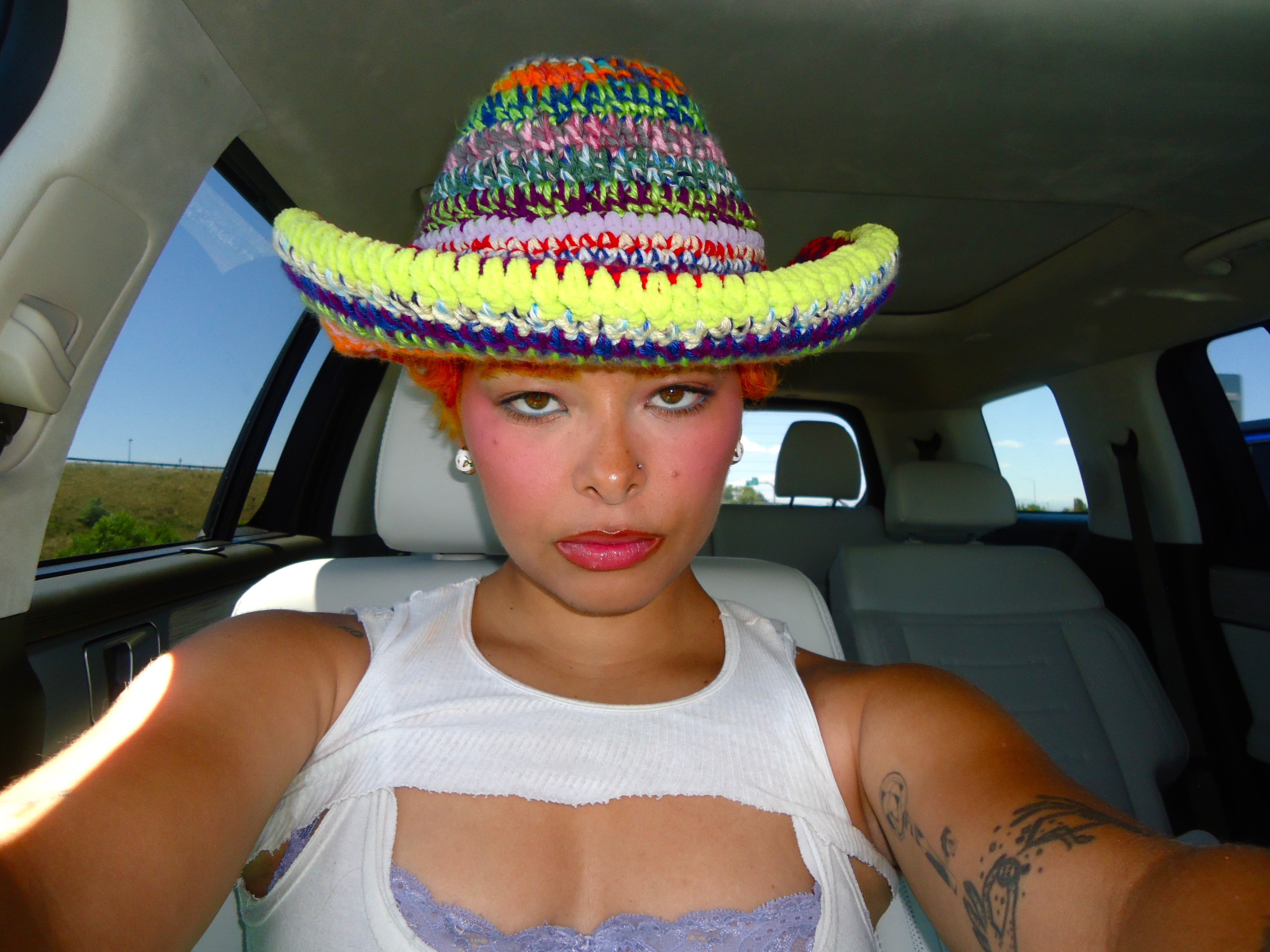 Young woman wearing a colorful woven hat and white top inside a vehicle, with a serious expression.