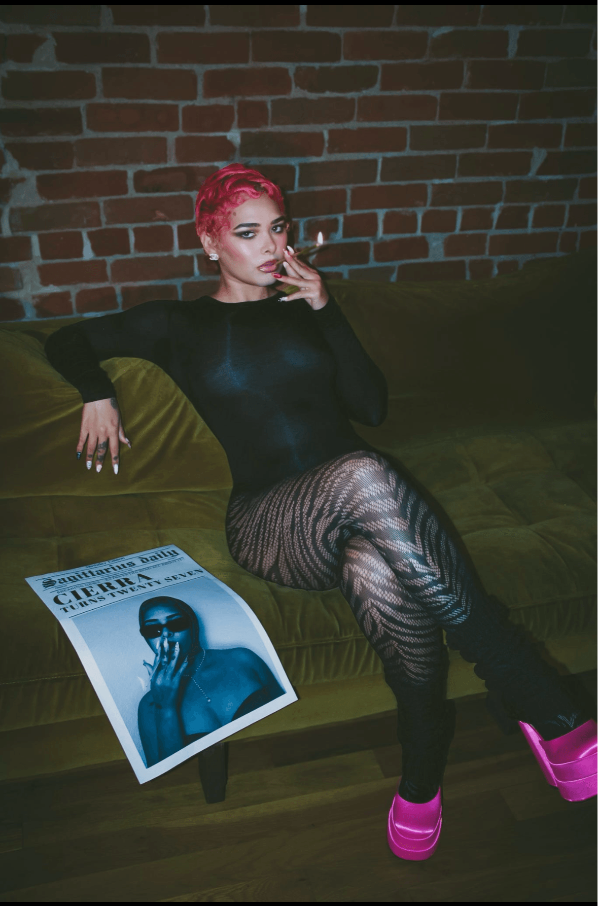 Woman with pink hair sitting on a green couch against a brick wall, wearing black outfit and pink shoes, with a magazine on the floor.