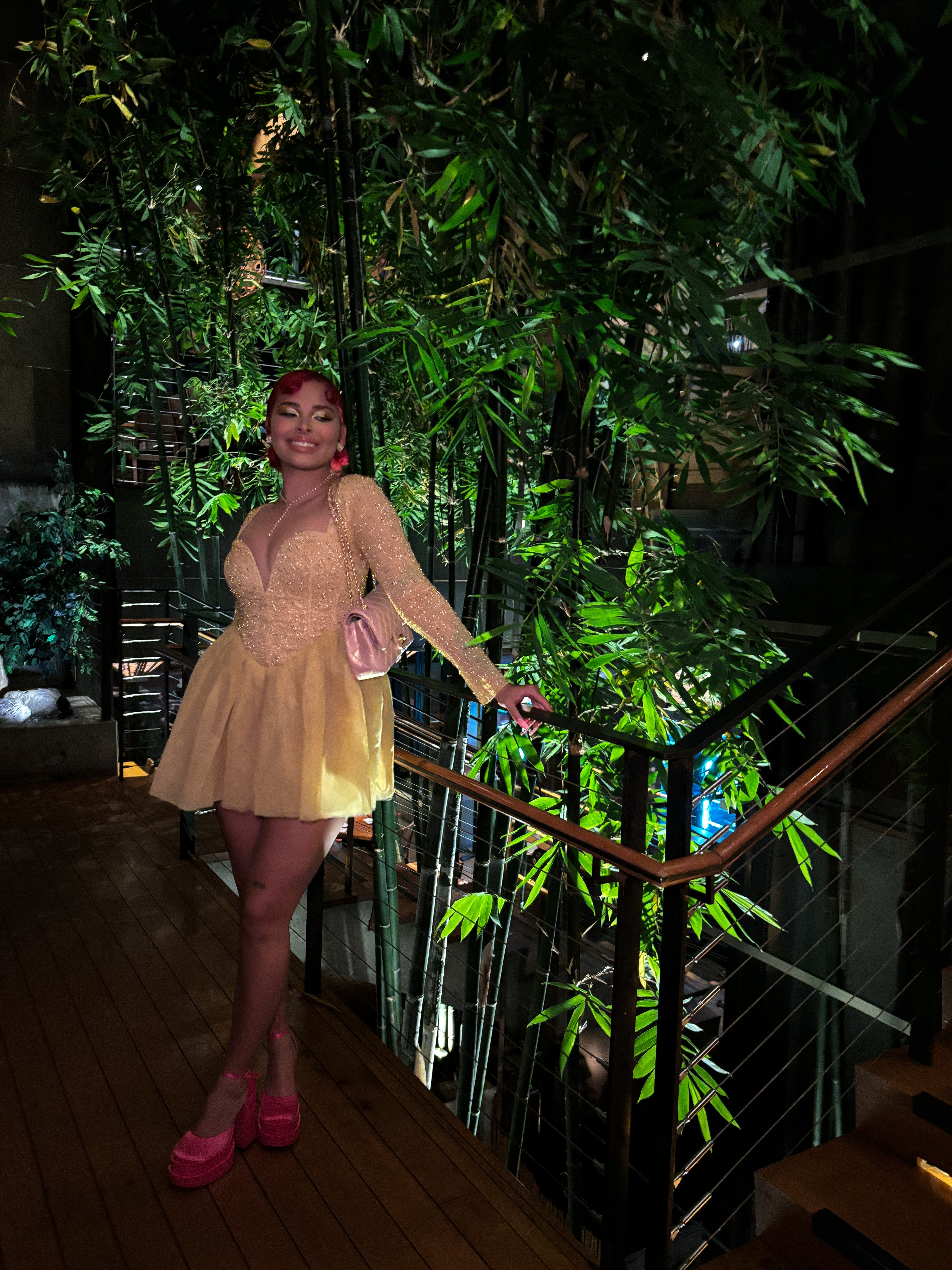 Woman in a beige dress and pink shoes standing on a wooden floor next to a railing, surrounded by green plants.