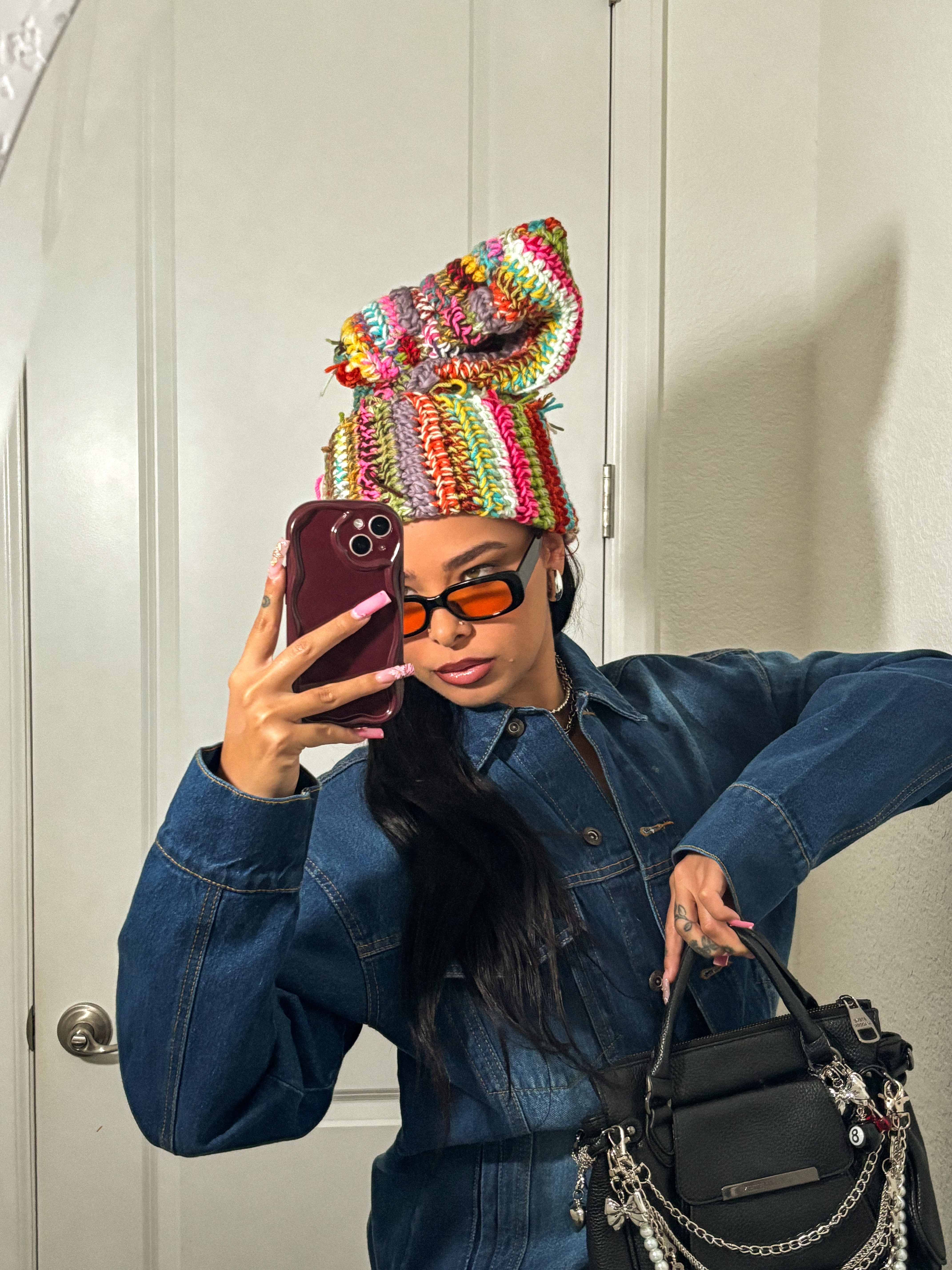 Person taking a mirror selfie wearing a colorful knitted hat, sunglasses, denim jacket, and holding a black bag.