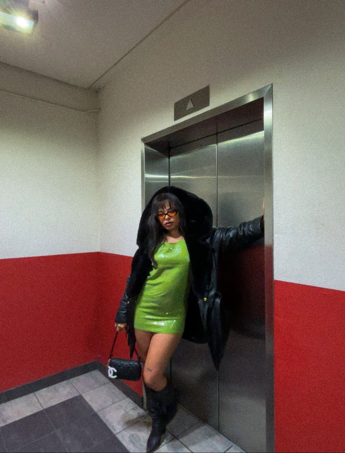 Woman in a green dress and black jacket standing in front of an elevator, holding a purse, with a red and white wall behind her.