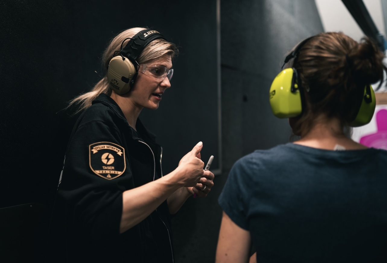 Two women wearing headphones and talking, one with glasses and a black jacket, the other with a bun and blue shirt.