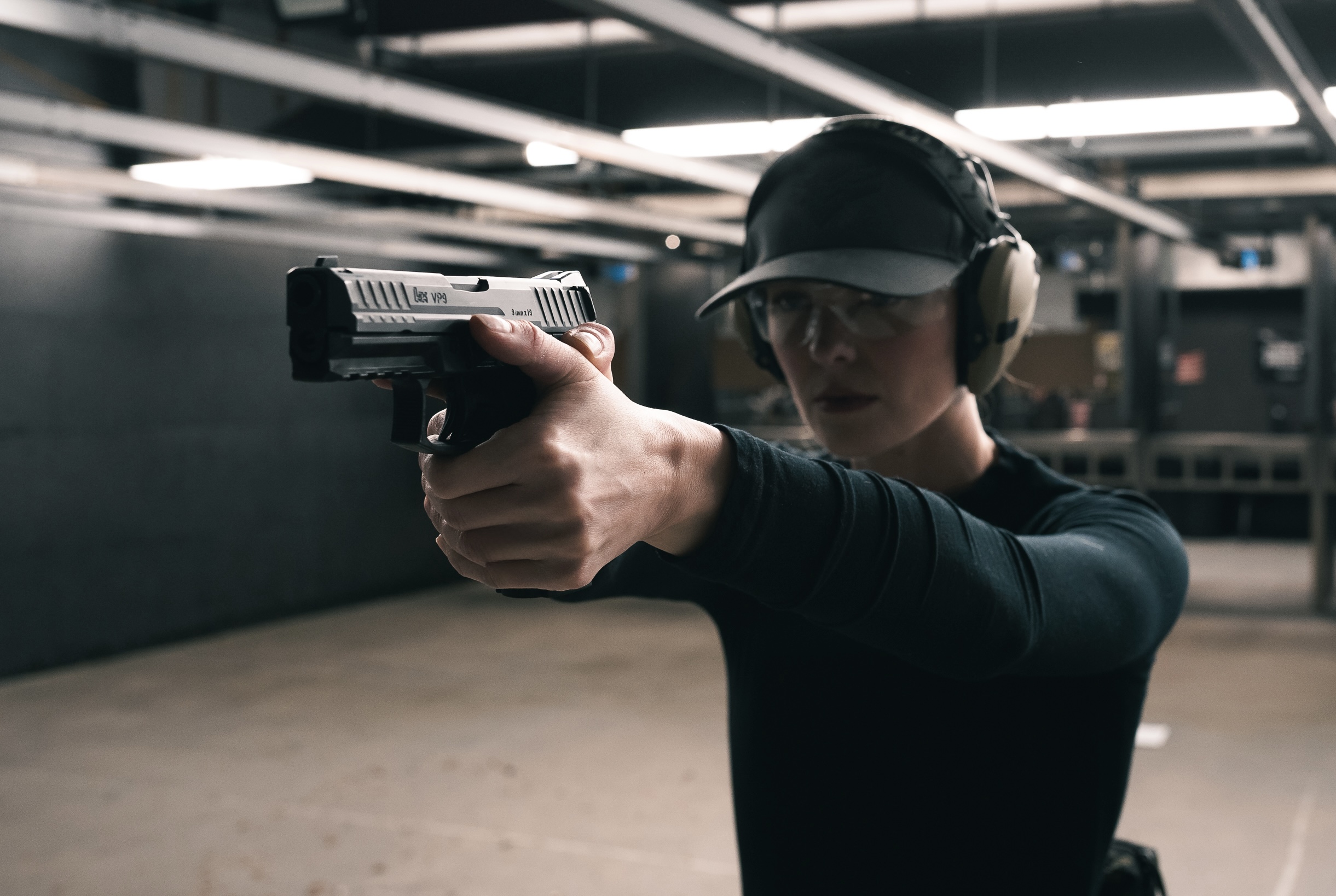 Person aiming a handgun at a shooting range, wearing ear protection and a cap, in an indoor setting.