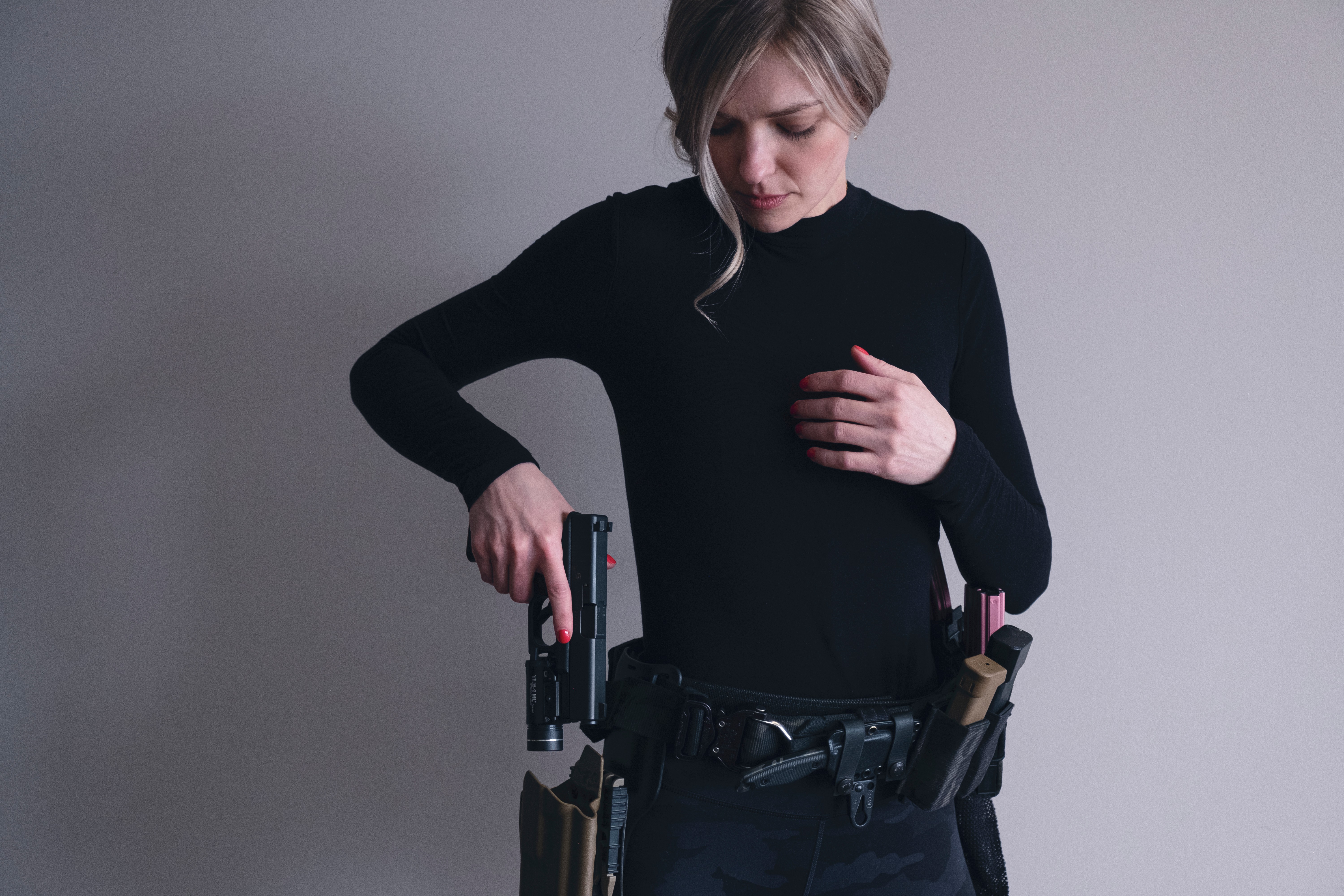 Person with short hair wearing a black long-sleeve shirt, holding a gun, with a belt holding a magazine and other items.