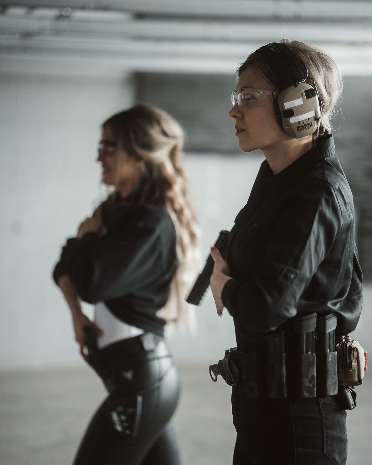 Two women in black uniforms, one with headphones and a gun, standing in an industrial setting.