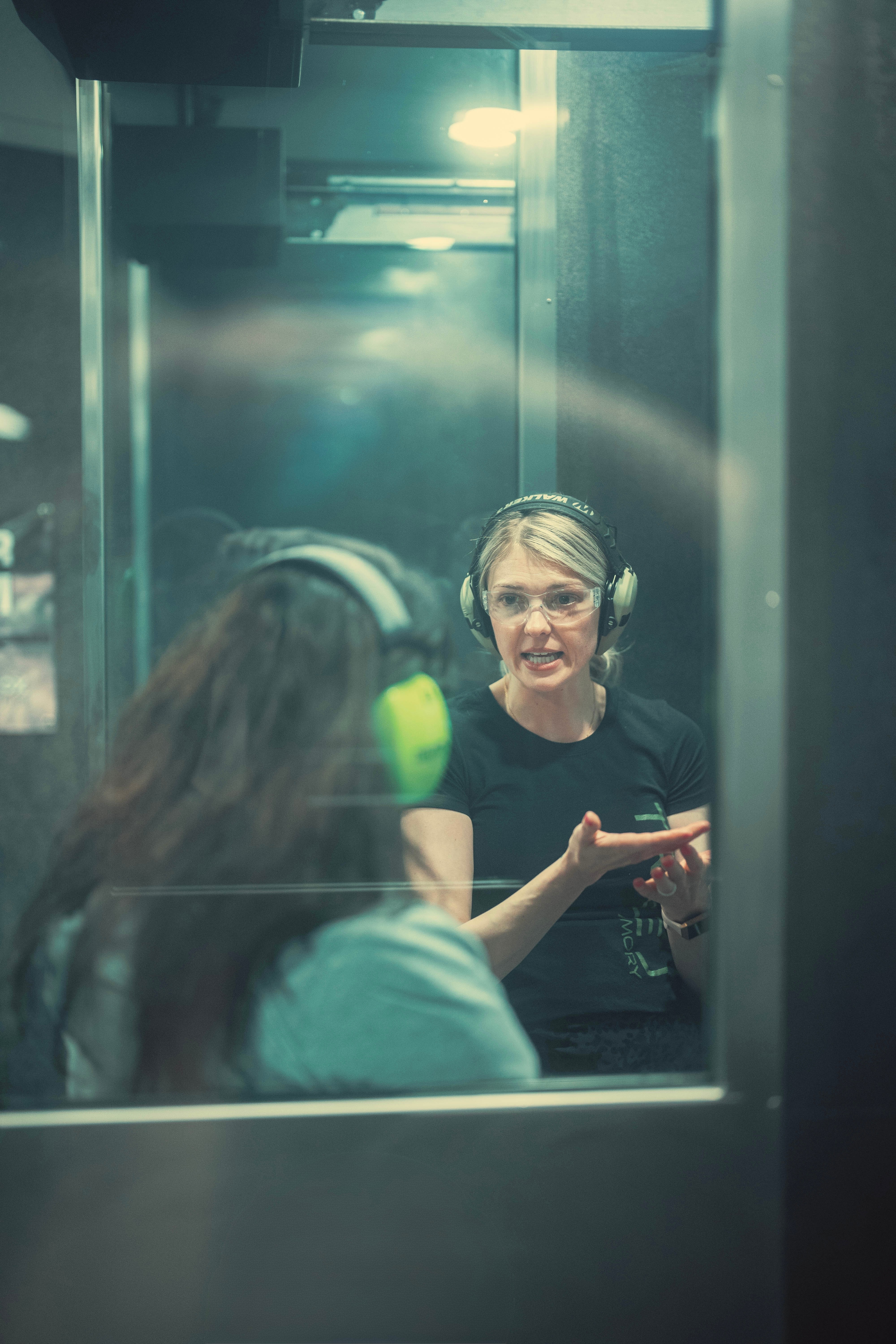 Two women in a recording studio, one wearing headphones and speaking, the other listening with headphones, reflected in a glass window.