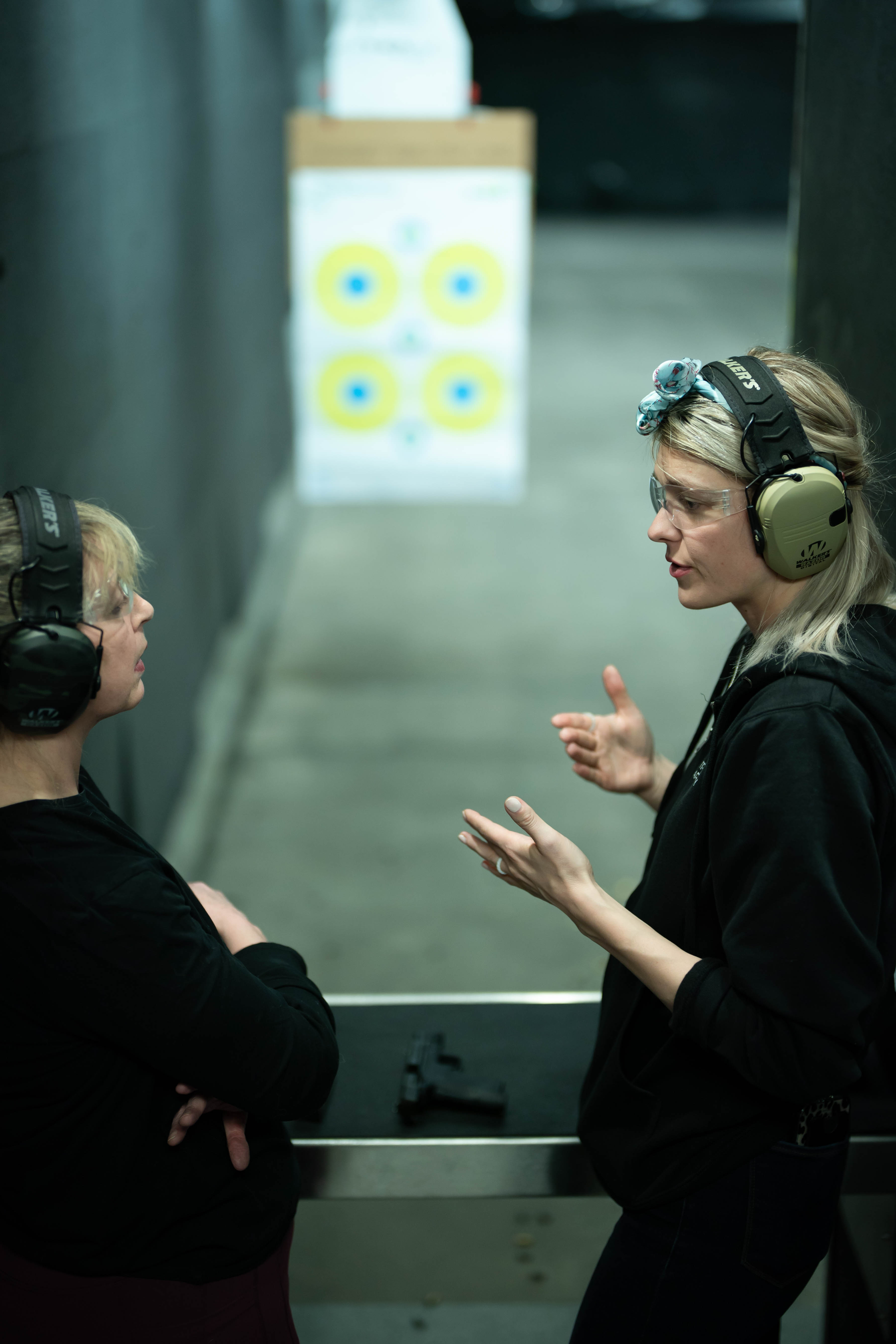 Two women with headphones engaged in conversation in an indoor shooting range, targets visible in the background.
