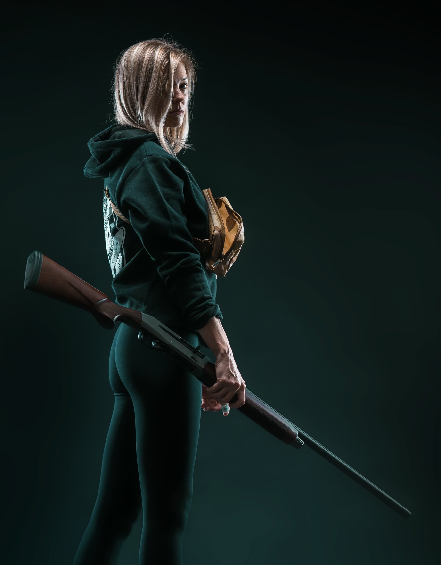 Woman holding a rifle, standing sideways, wearing a hoodie and leggings, with a dark background.